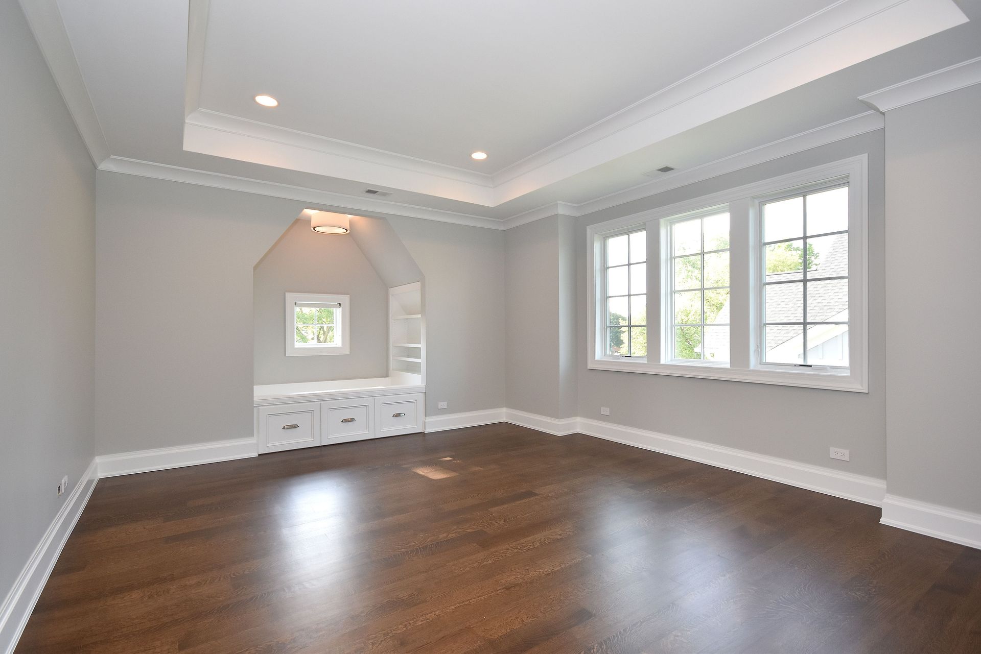 Empty living room with dark hardwood floors, white walls, tray ceiling, and large windows.
