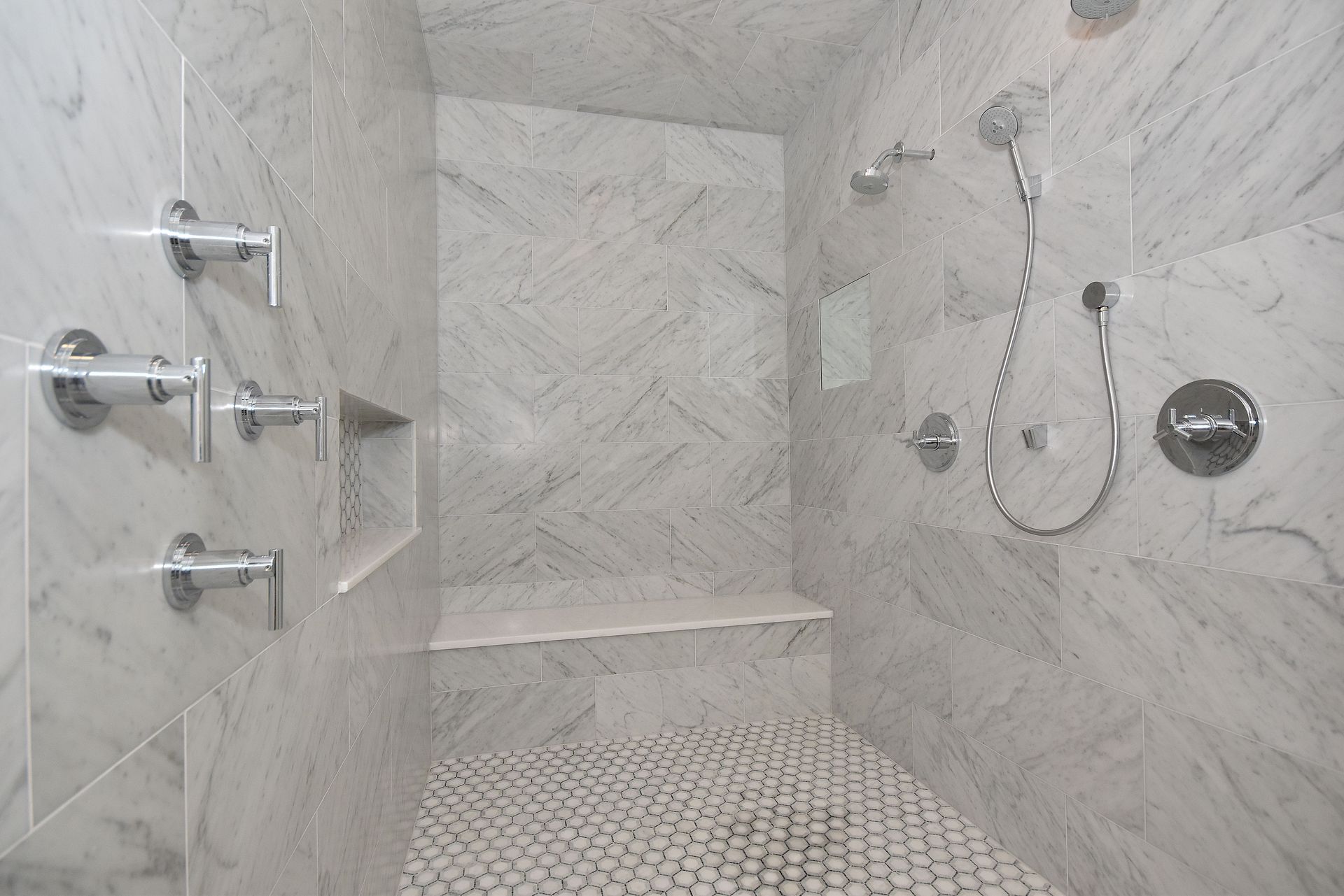 Modern marble-tiled shower with multiple chrome fixtures, built-in shelf, and pebble mosaic floor.