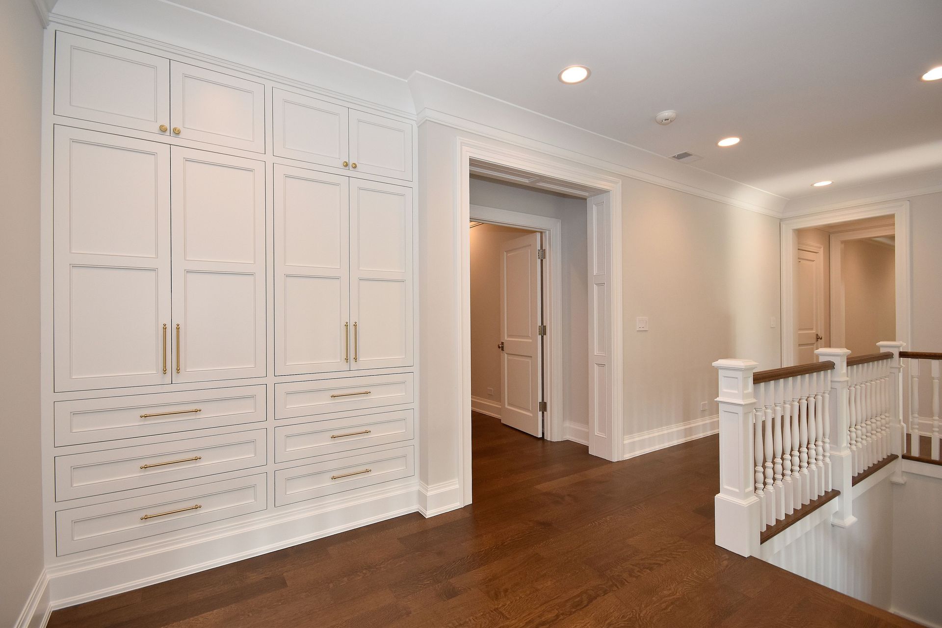 Bright white hallway with built-in cabinets, wood floors, recessed lights, and a white stair railing
