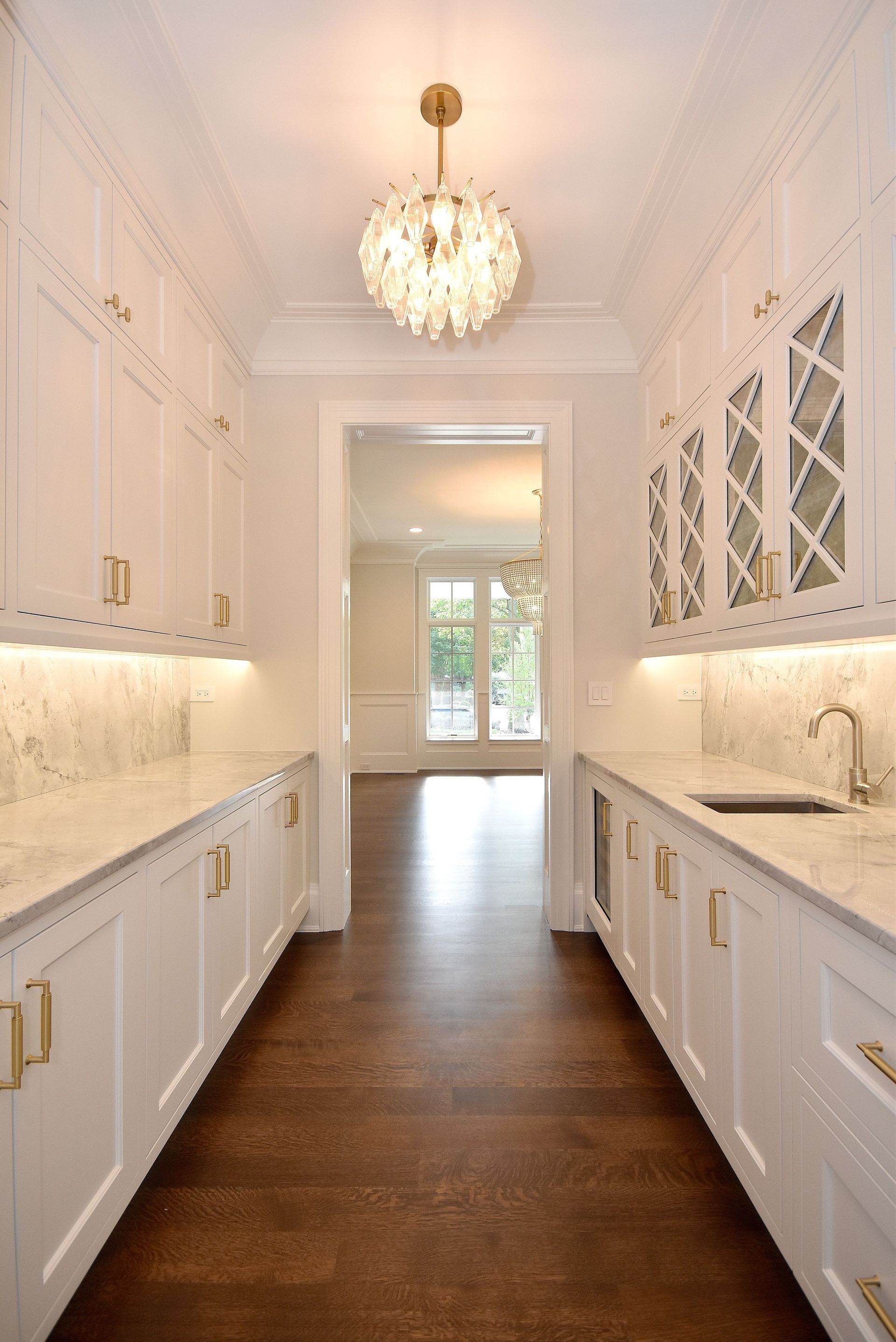 Bright white galley kitchen with marble counters, gold fixtures, and a chandelier over dark wood flooring