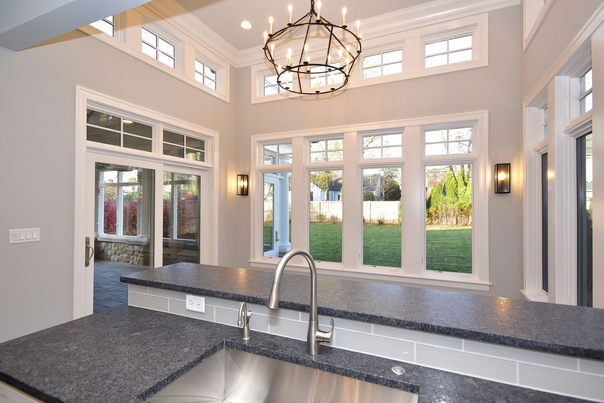 Bright kitchen with large windows, granite counters, sink, and chandelier overlooking a backyard