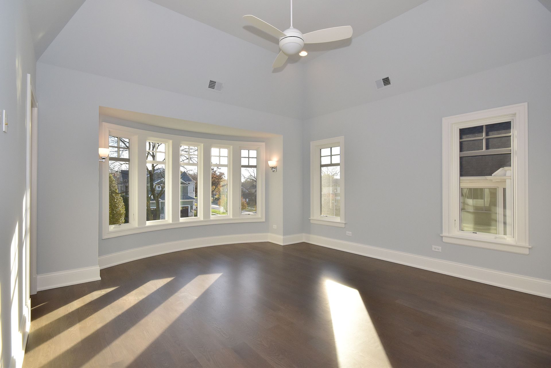 Empty bright living room with dark hardwood floors, bay windows, and a ceiling fan