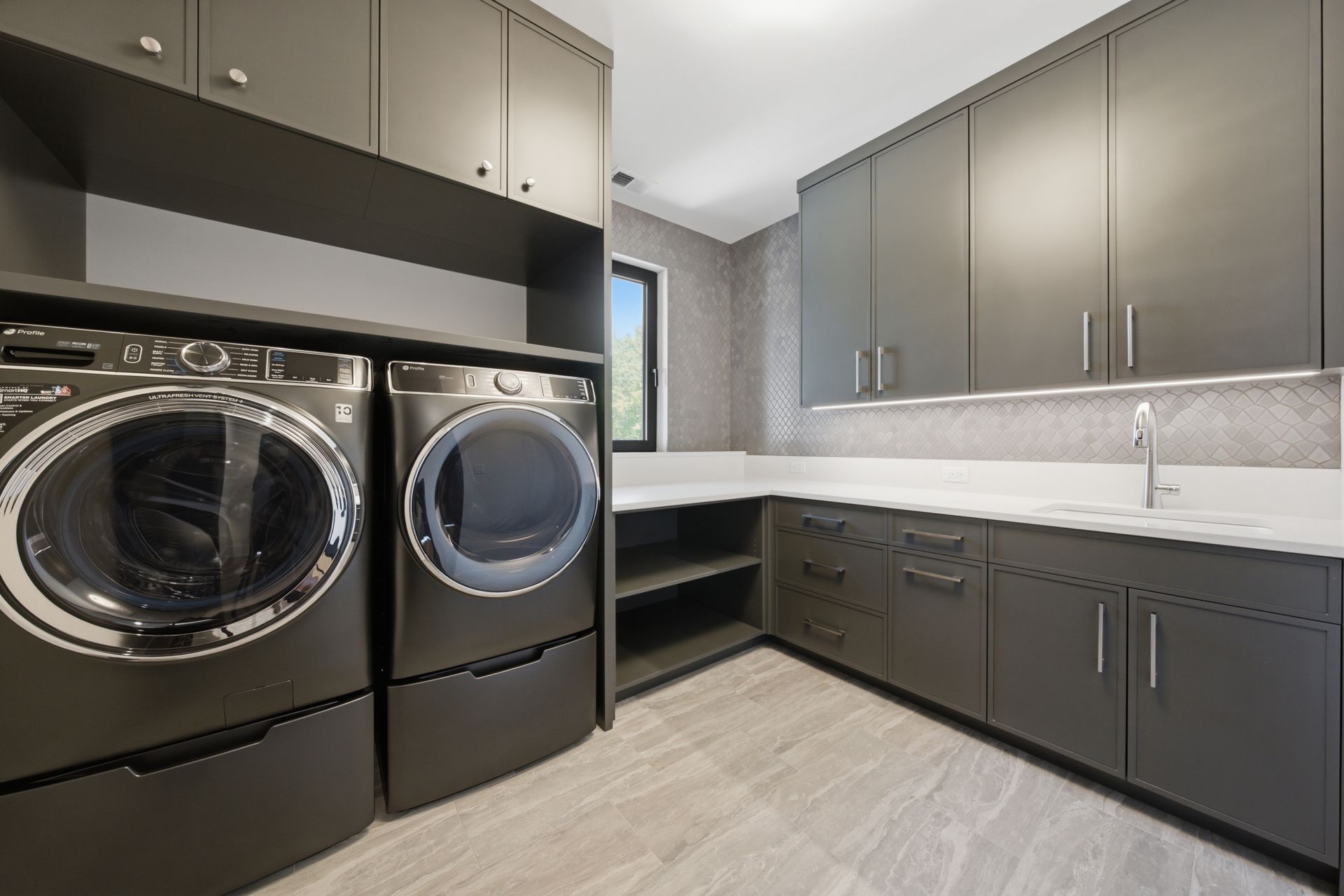 Modern laundry room with black washer and dryer, gray cabinets, and a long countertop by a window