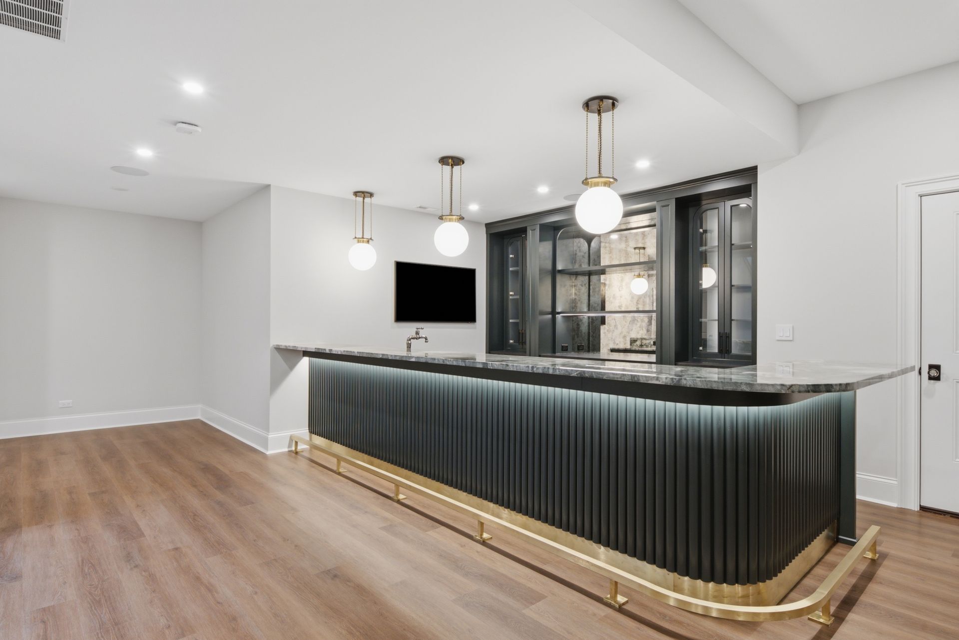 Modern home bar with dark ribbed front, gold trim, pendant lights, and light wood floors