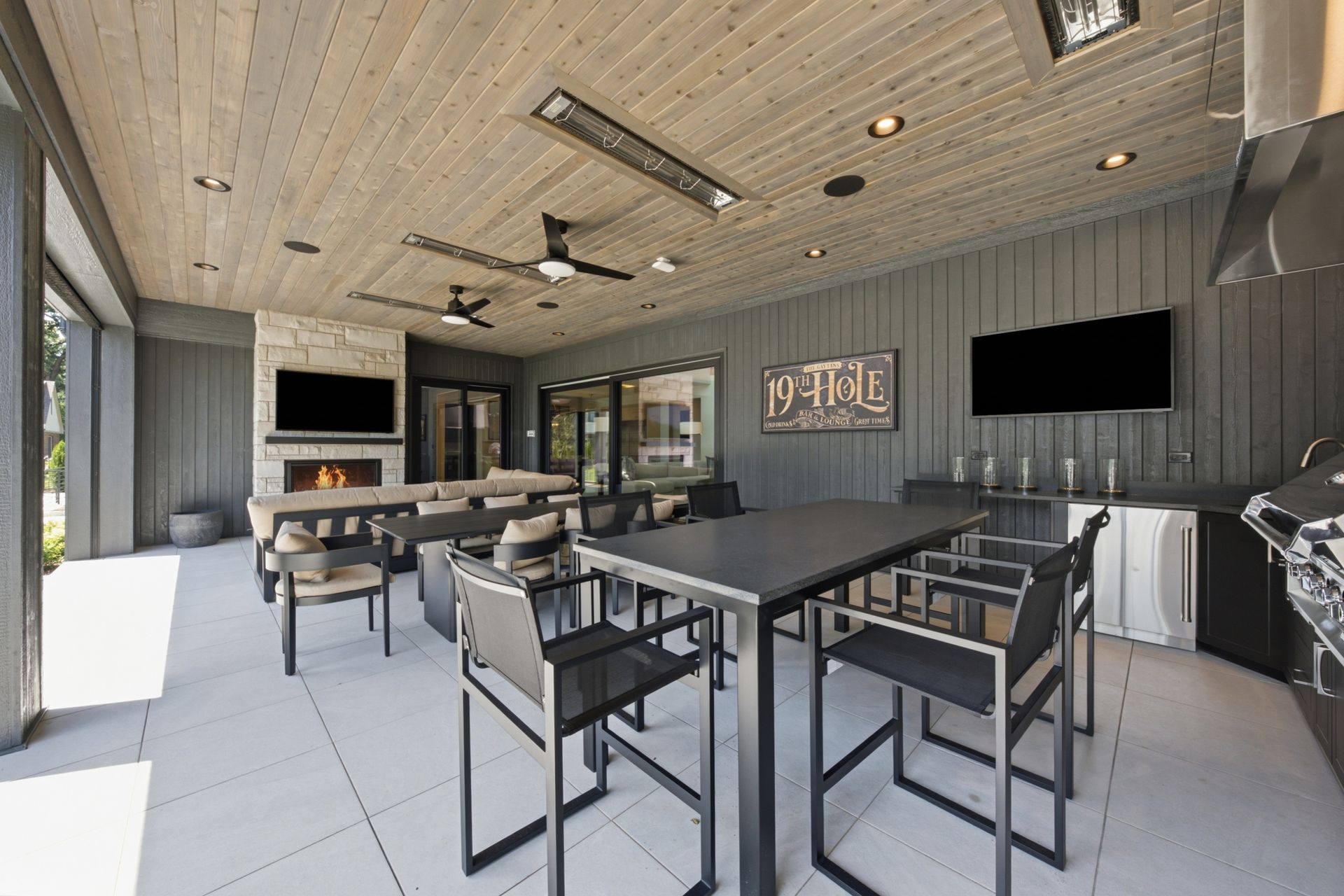 Modern covered patio with bar seating, large fireplace, ceiling fans, and wall-mounted TVs