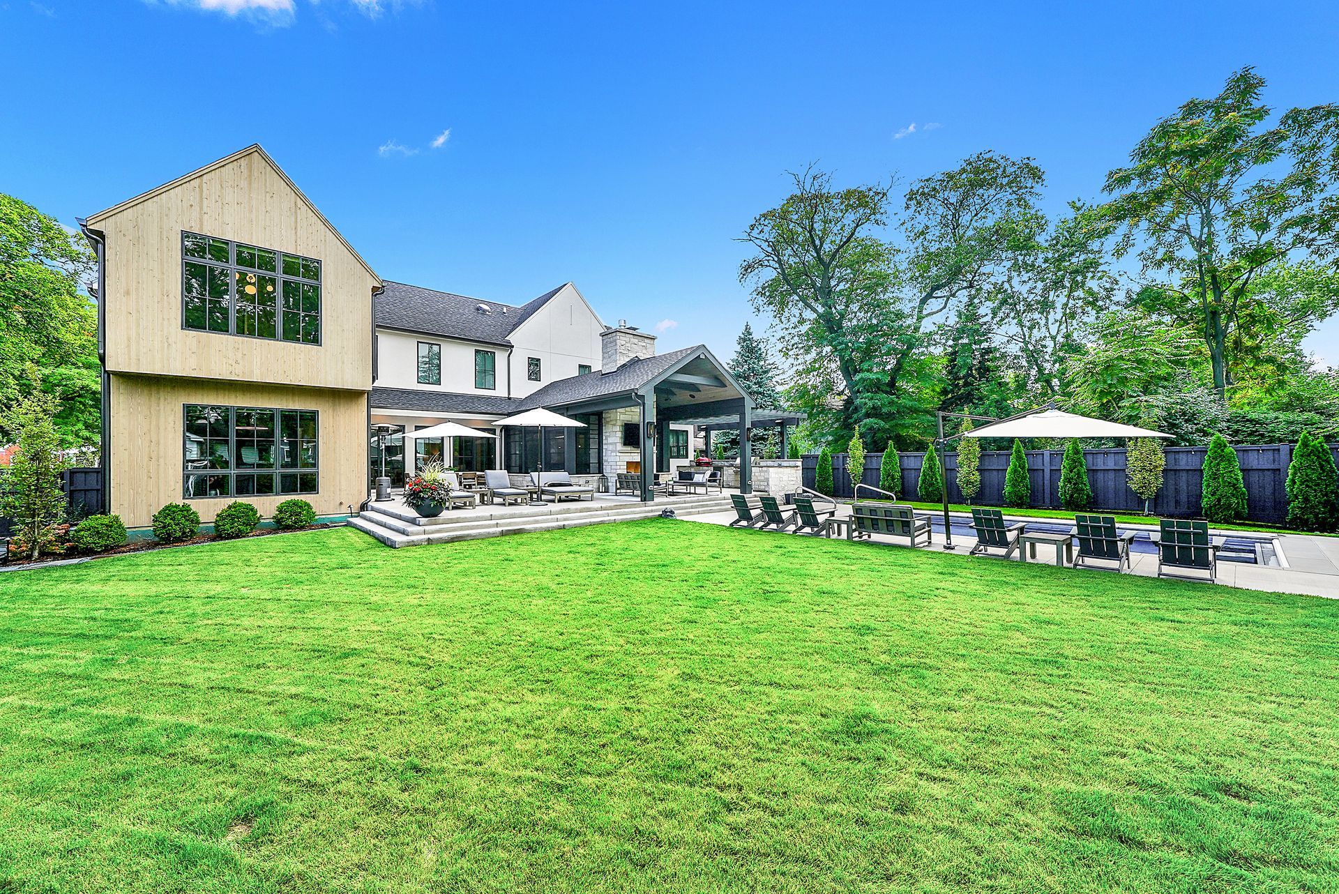 Spacious backyard with green lawn, modern house, patio seating, and trees under a bright blue sky