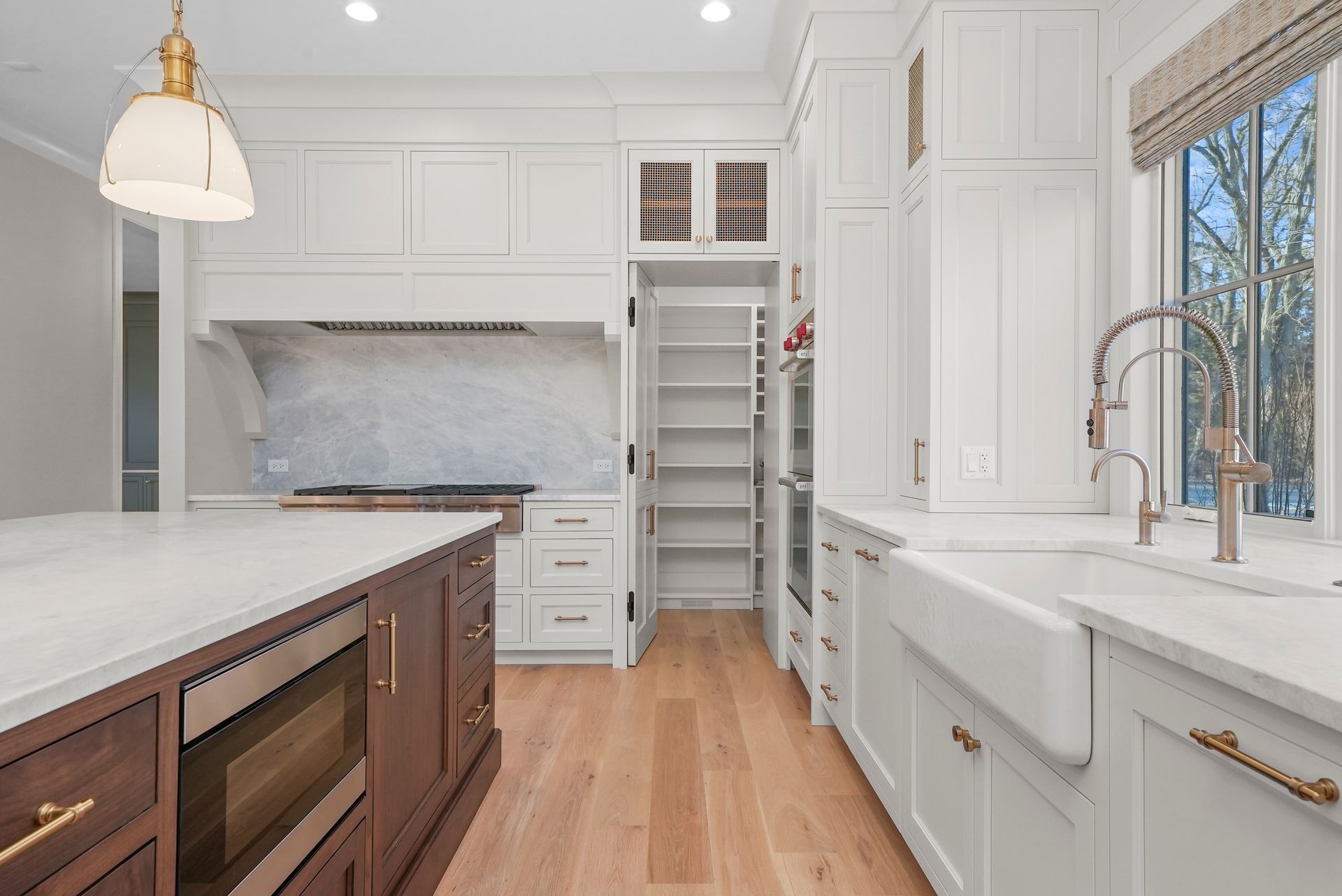 Bright white kitchen with marble island, wood floor, stainless range, and large window over farmhouse sink