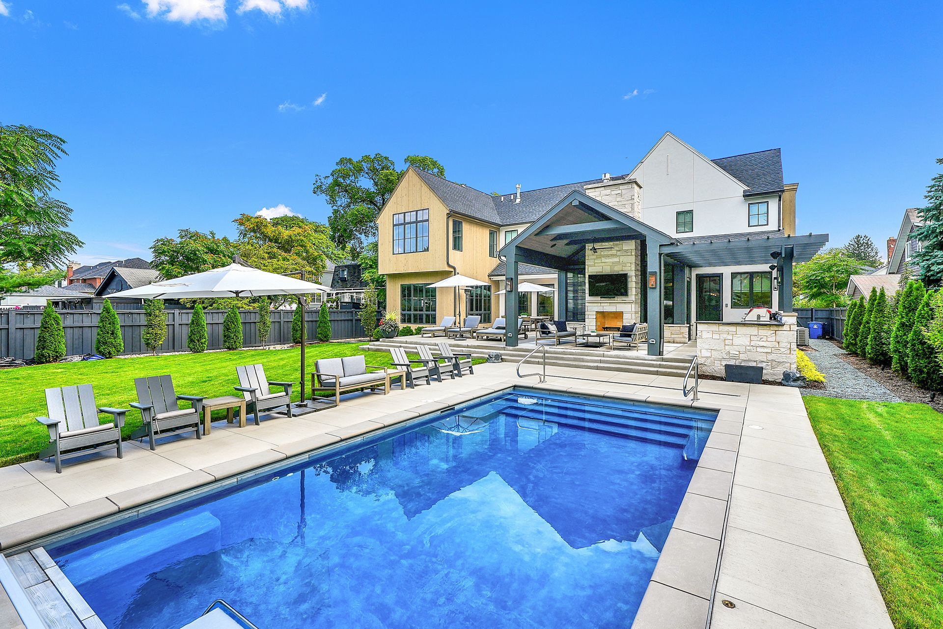 Modern backyard with a blue swimming pool, patio seating, and a large house under a sunny sky