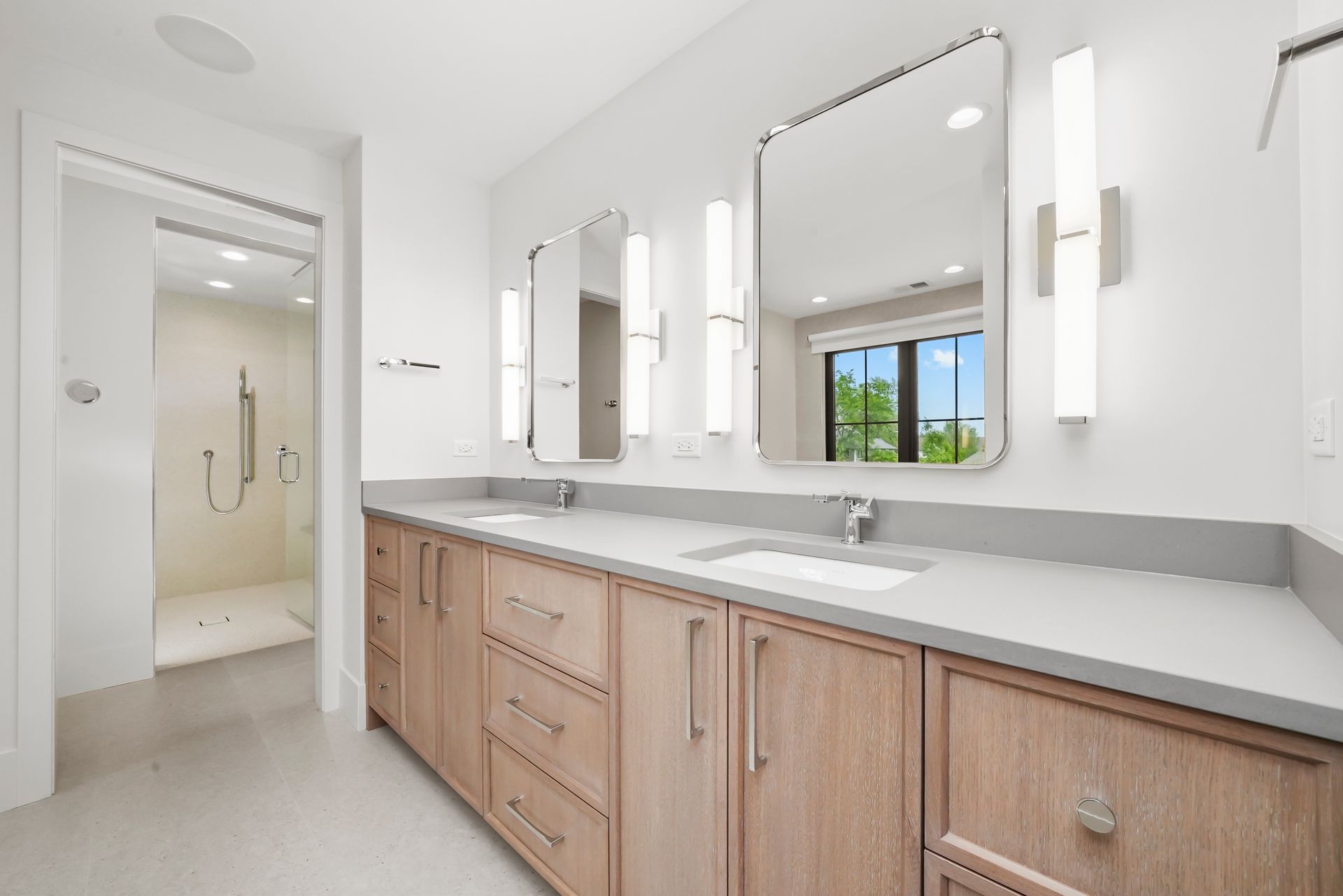 Bright bathroom vanity with wood cabinets, large mirror, and walk-in shower visible through open doorway.