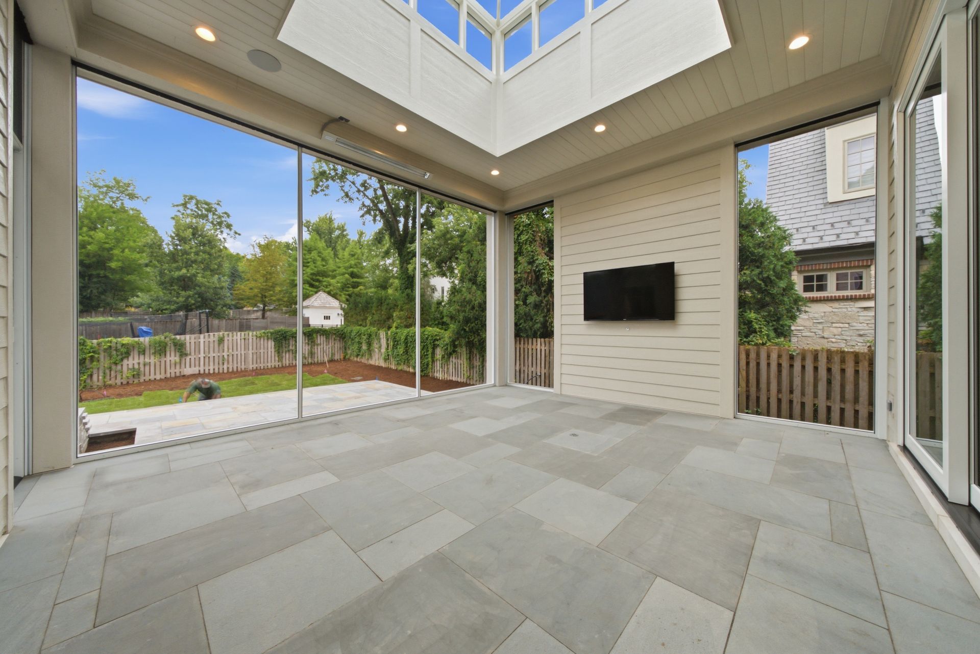 Modern covered patio with large sliding glass doors, tiled floor, and a wall-mounted TV overlooking a backyard
