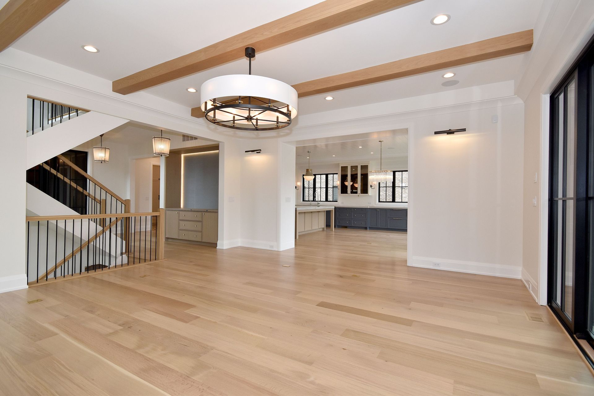 Spacious modern living room with light wood floors, white walls, chandelier, and open view to kitchen.