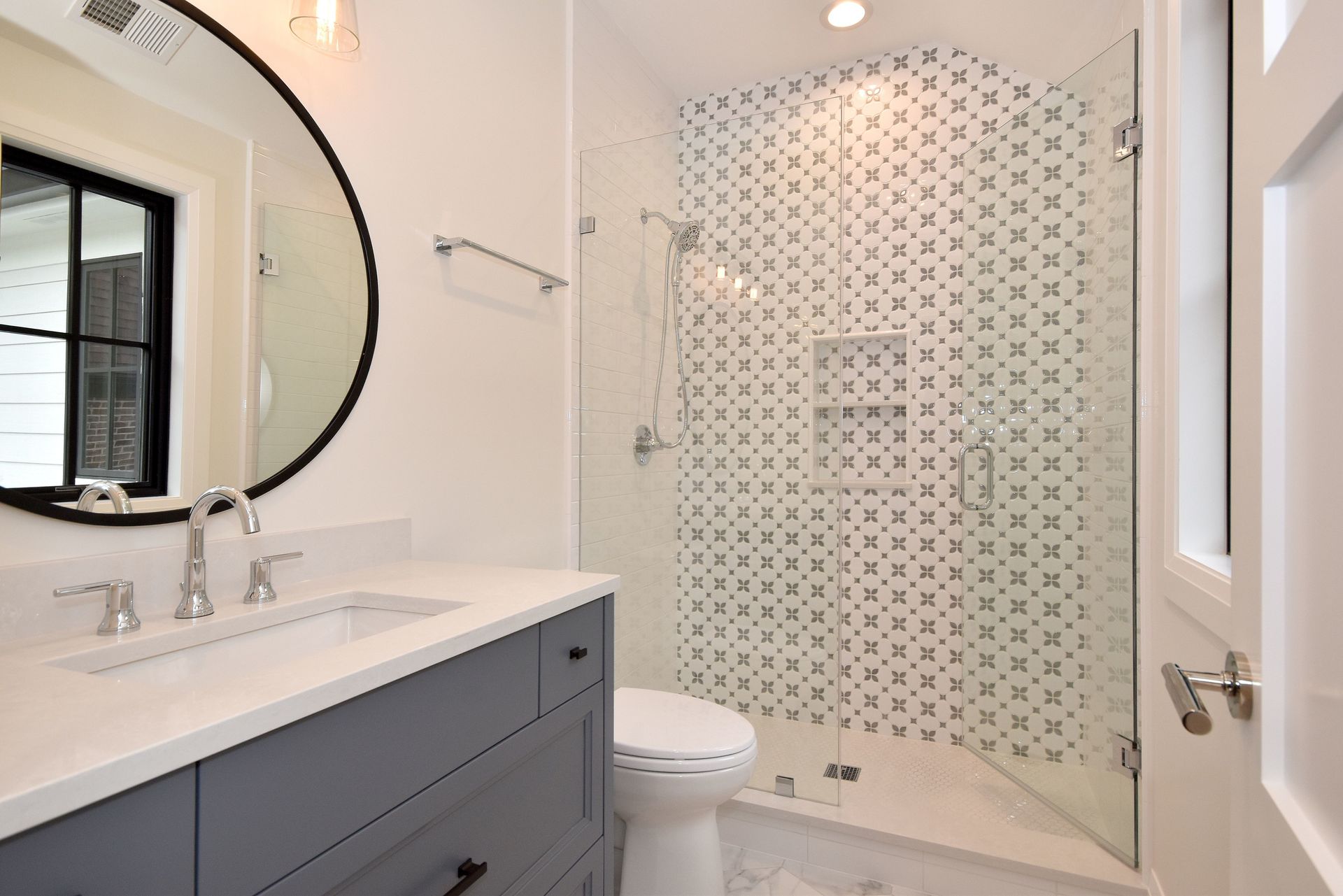 Bright modern bathroom with vanity, round mirror, toilet, and glass shower with patterned tile.