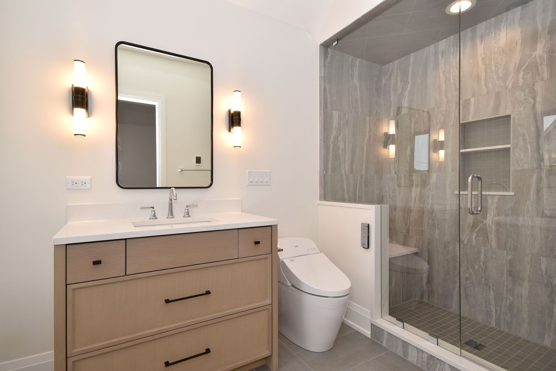 Modern bathroom with beige vanity, mirror, toilet, and glass shower tiled in gray.