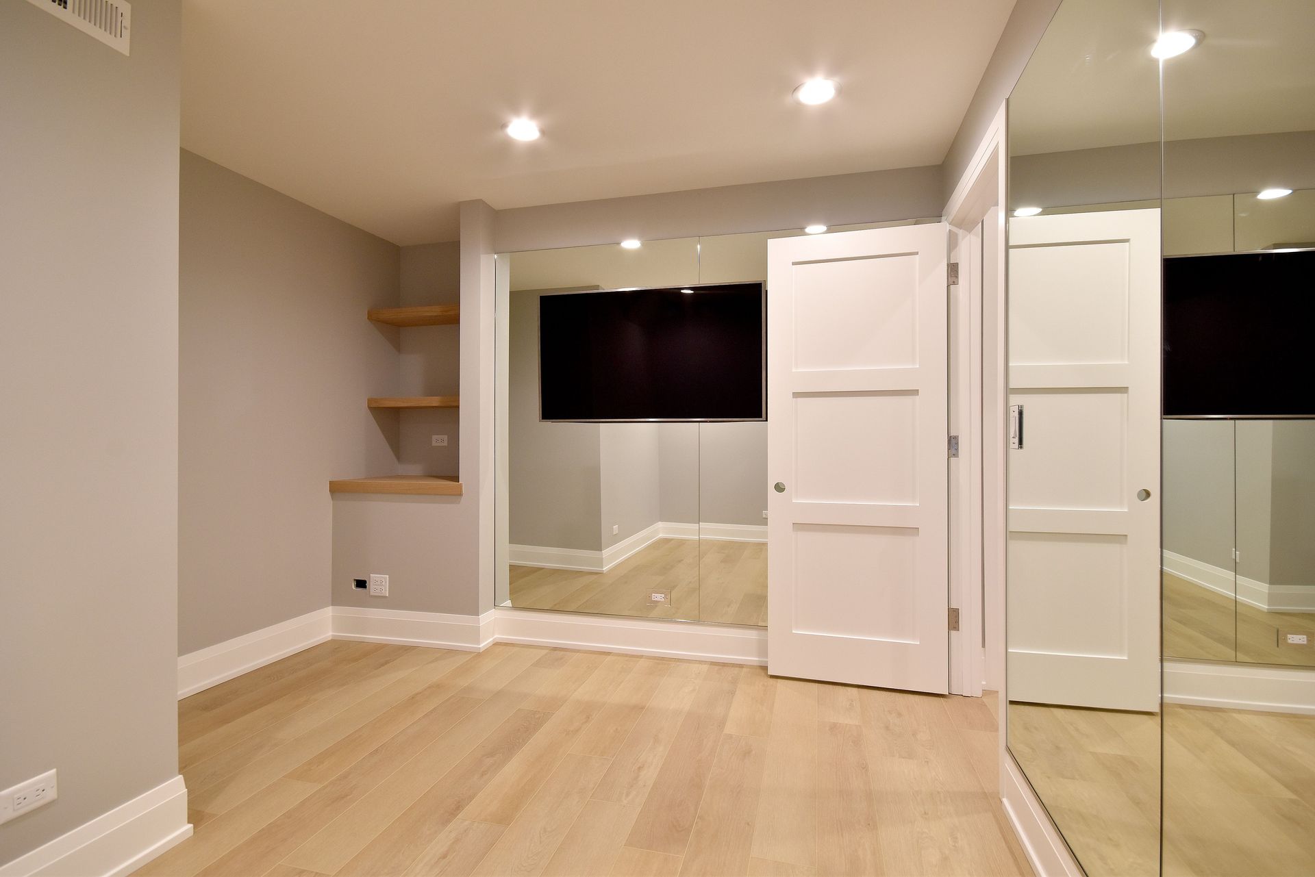 Empty beige bedroom with light wood floors, mirrored closet doors, and a wall-mounted TV.