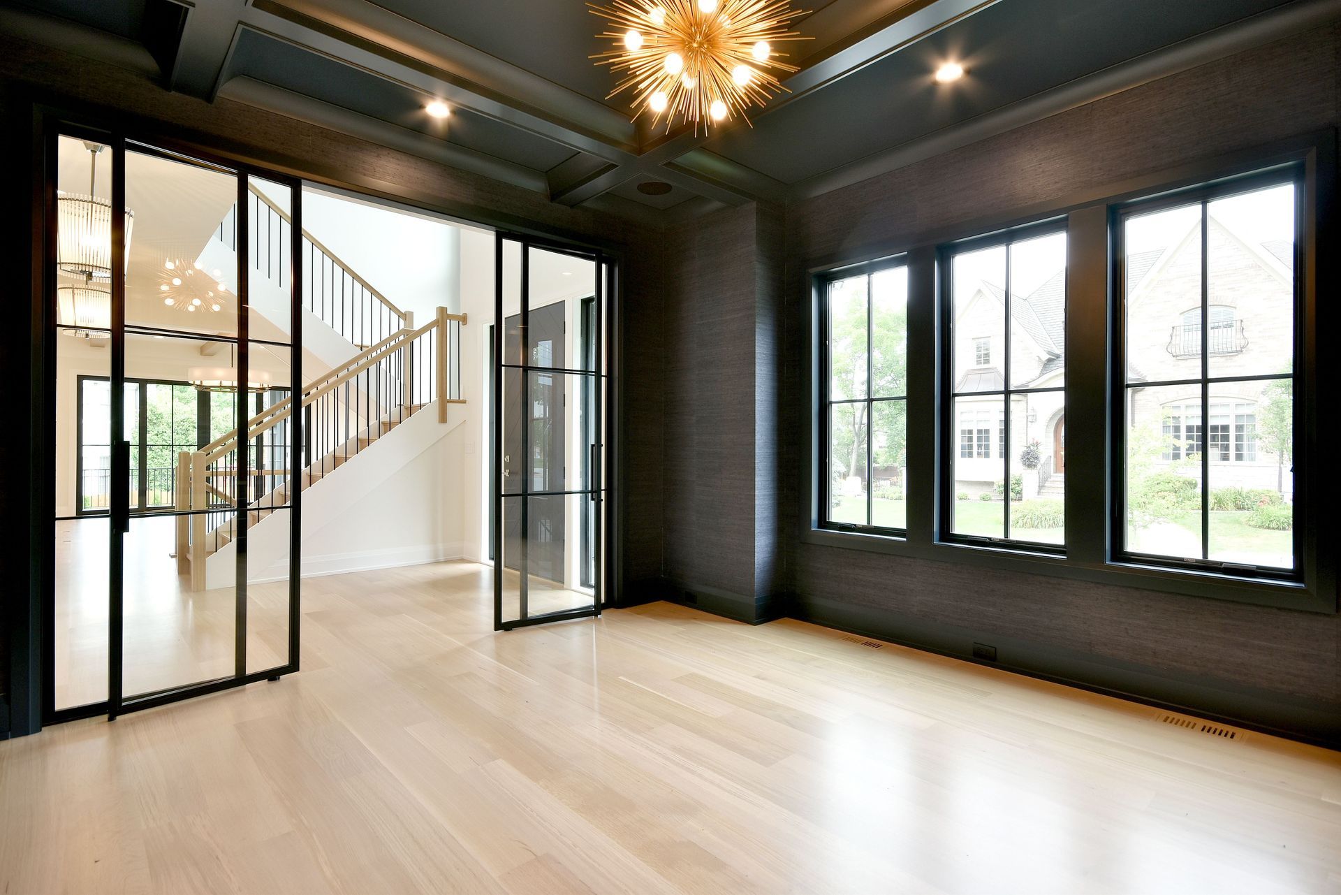 Modern empty living room with black-framed windows, light wood floors, and a staircase visible through open doors