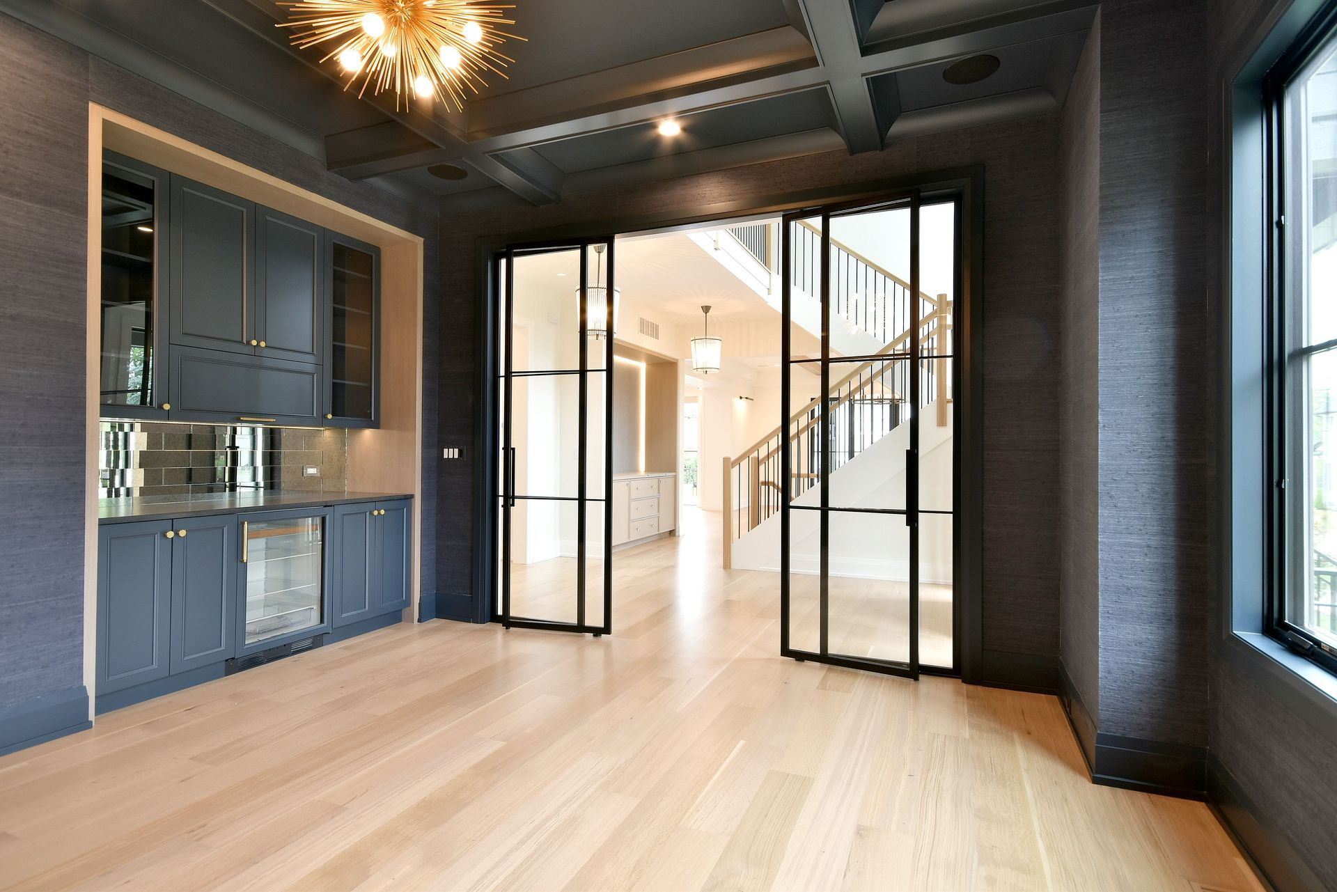 Modern empty room with light wood floors, dark walls, built-in cabinets, and glass doors to a staircase.