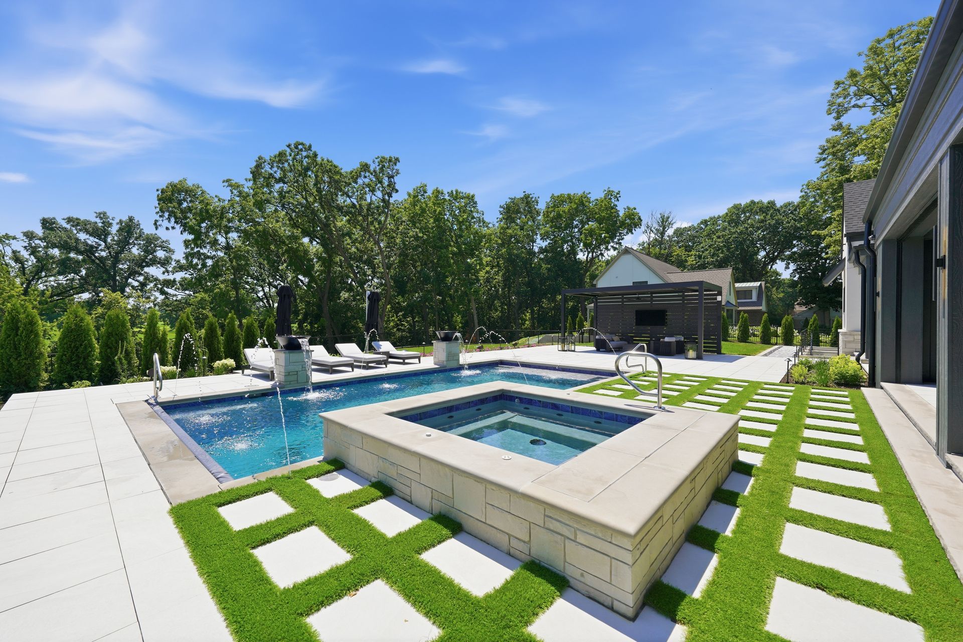 Modern backyard with pool, hot tub, and white pavers set in green grass under a sunny blue sky
