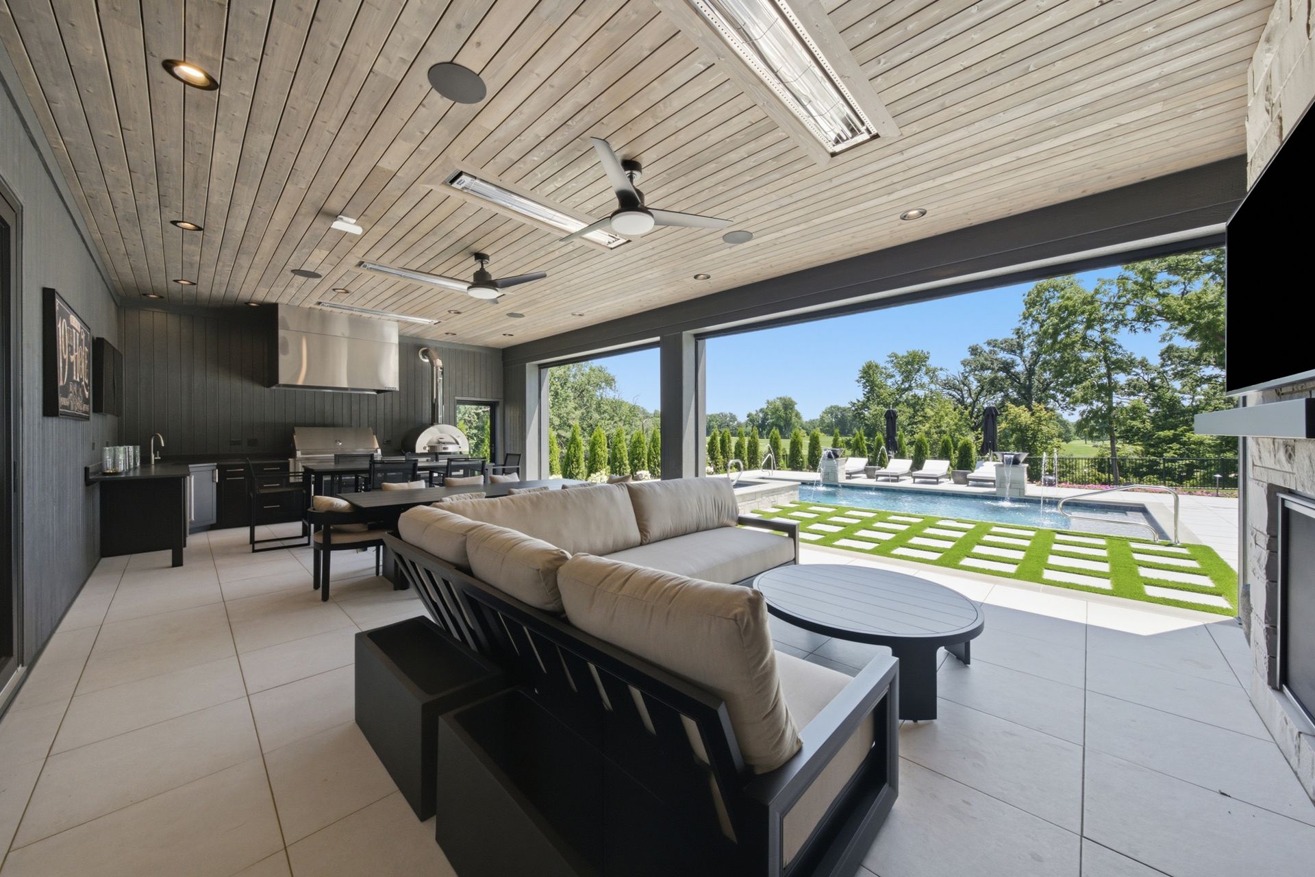 Modern covered patio with outdoor sofa, kitchen area, and open view of lawn and trees