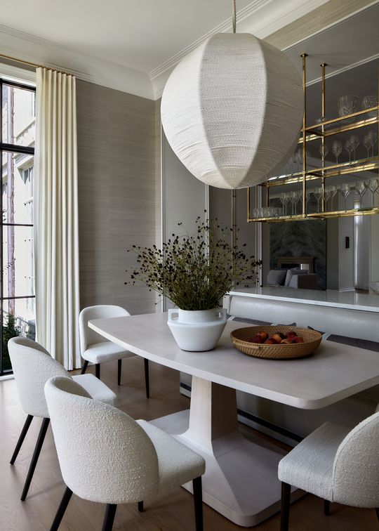 Elegant dining room with a white table, textured chairs, pendant light, and gold shelving by a window.