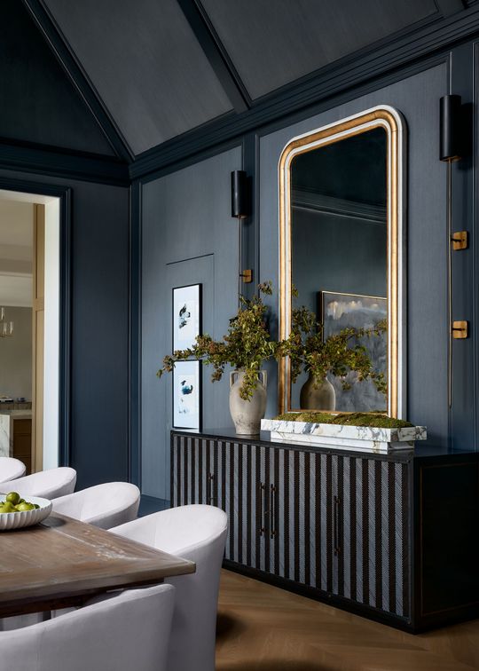 Elegant dining room with dark walls, a gold-framed mirror, and a striped black sideboard with flowers