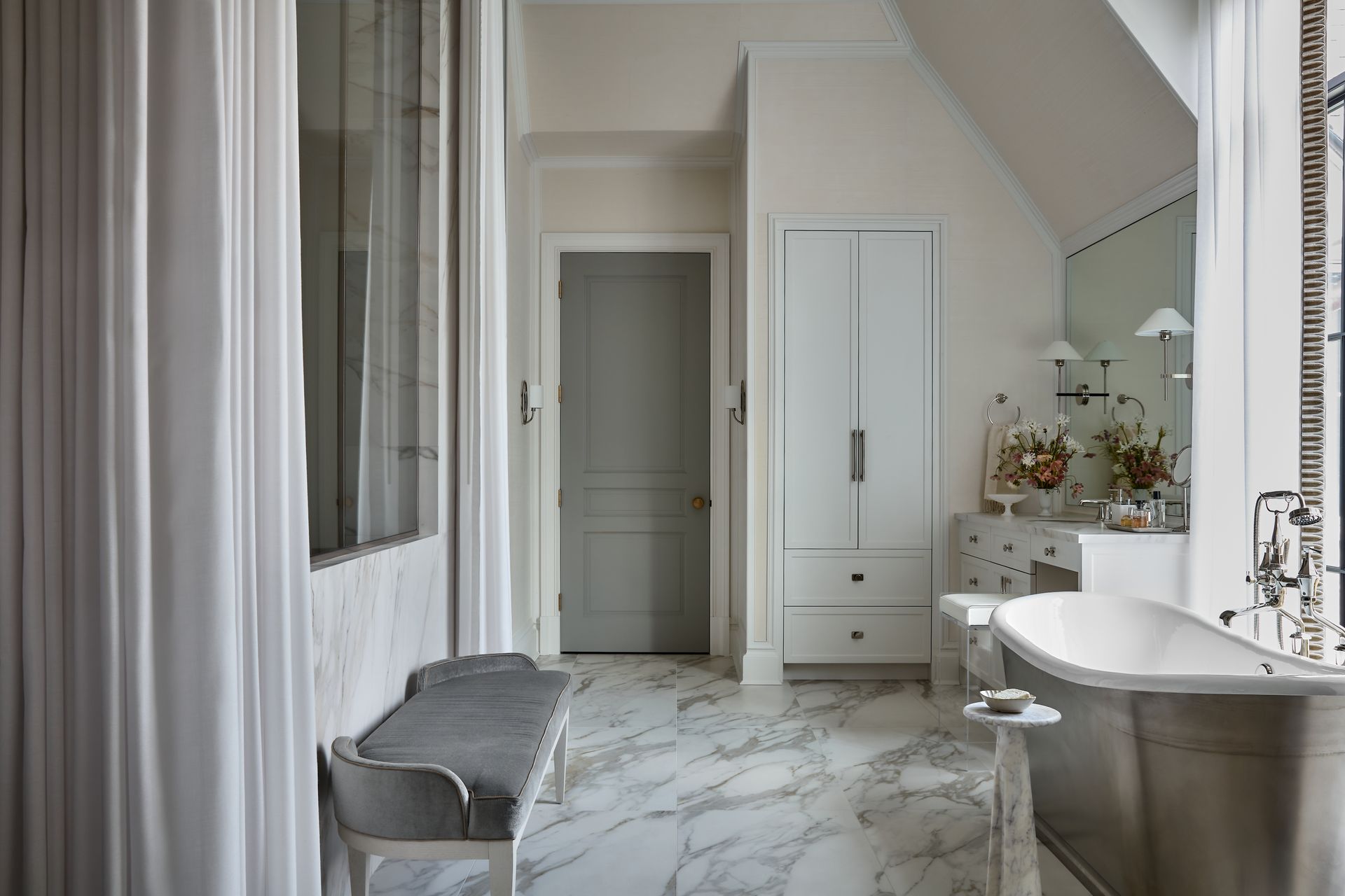 Elegant bathroom with marble floor, freestanding tub, gray bench, and white cabinetry in soft neutral tones