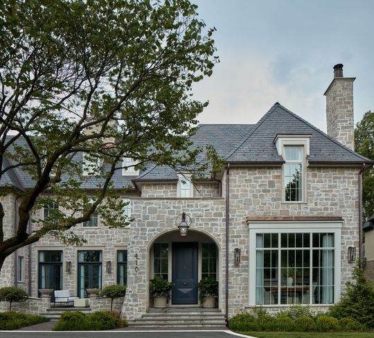 Stone house exterior with a slate roof, large windows, and a front entrance framed by trees