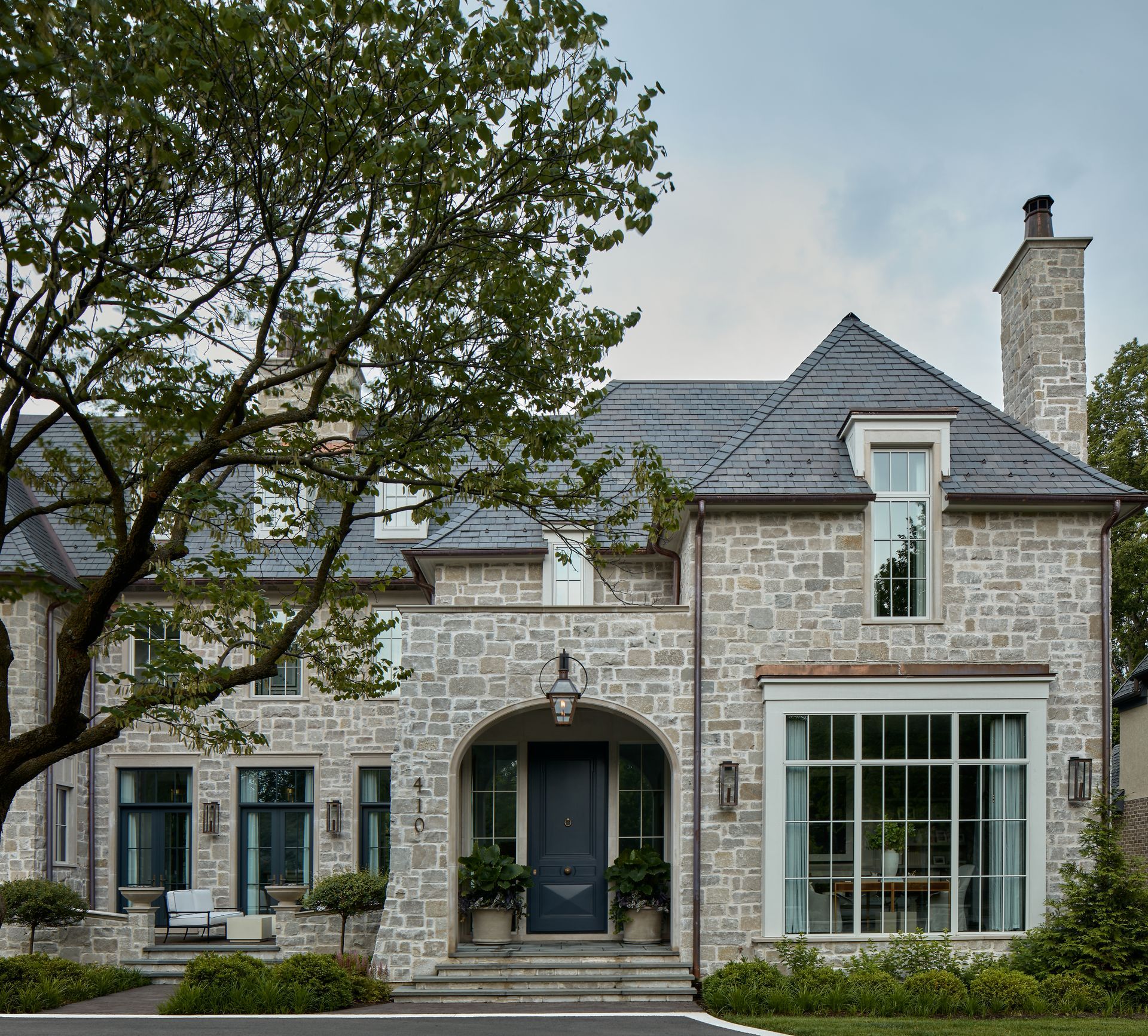 Stone house with gray roof, large bay window, and front steps, framed by a leafy tree and hedges.