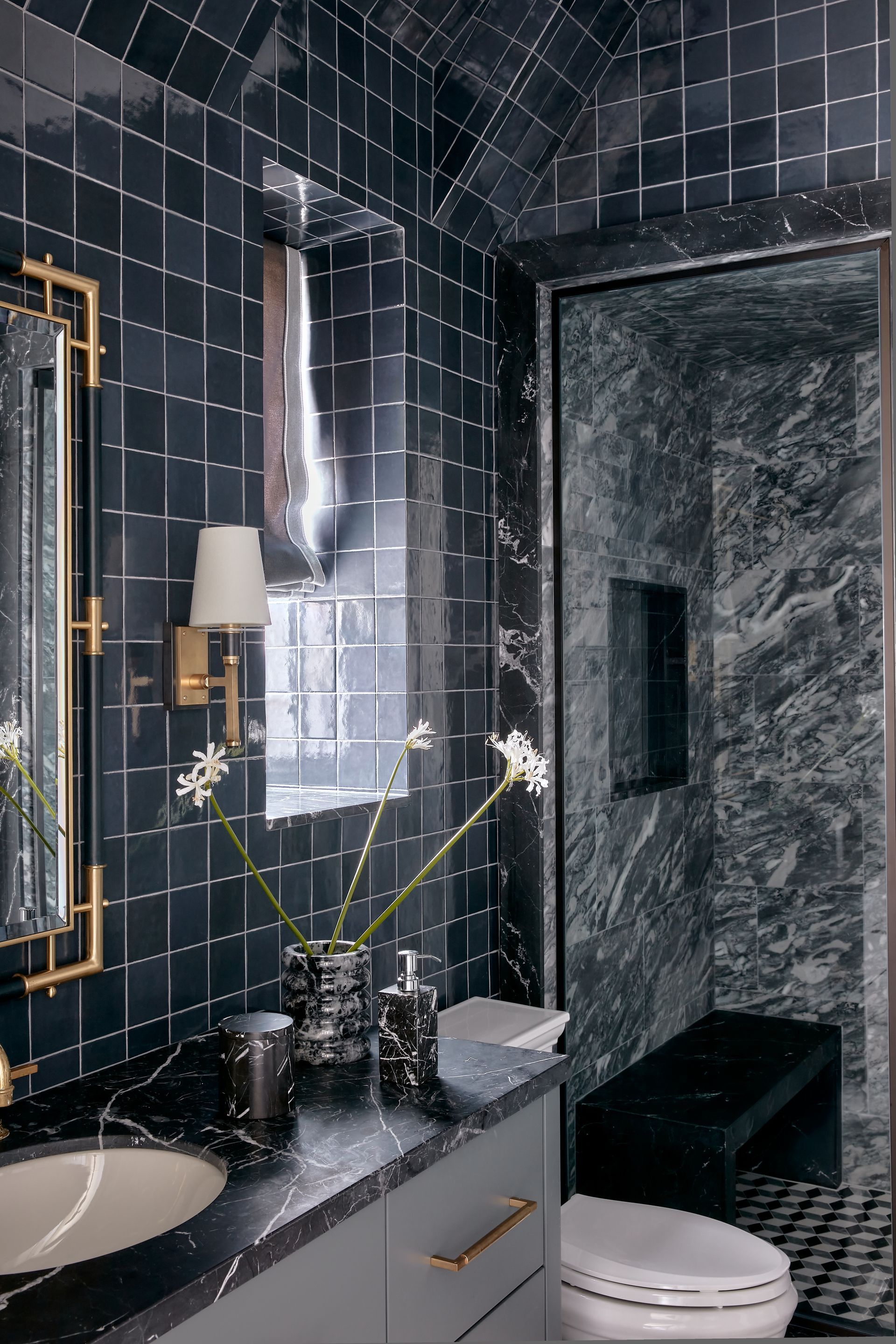 Dark modern bathroom with black tiled walls, marble shower, and a vanity with flowers and a wall mirror