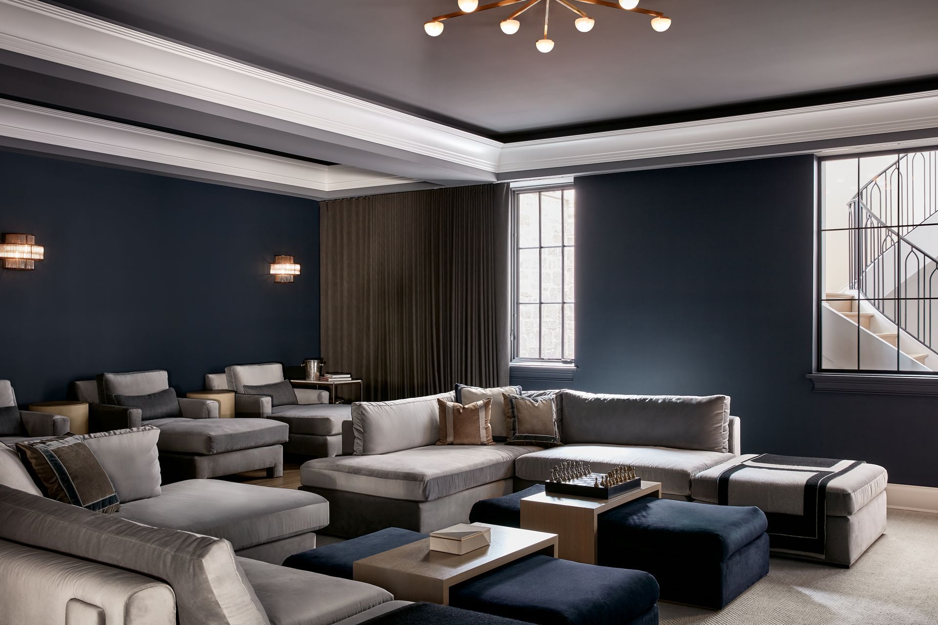Modern living room with gray sectional sofas, dark walls, and large windows with blackout shades