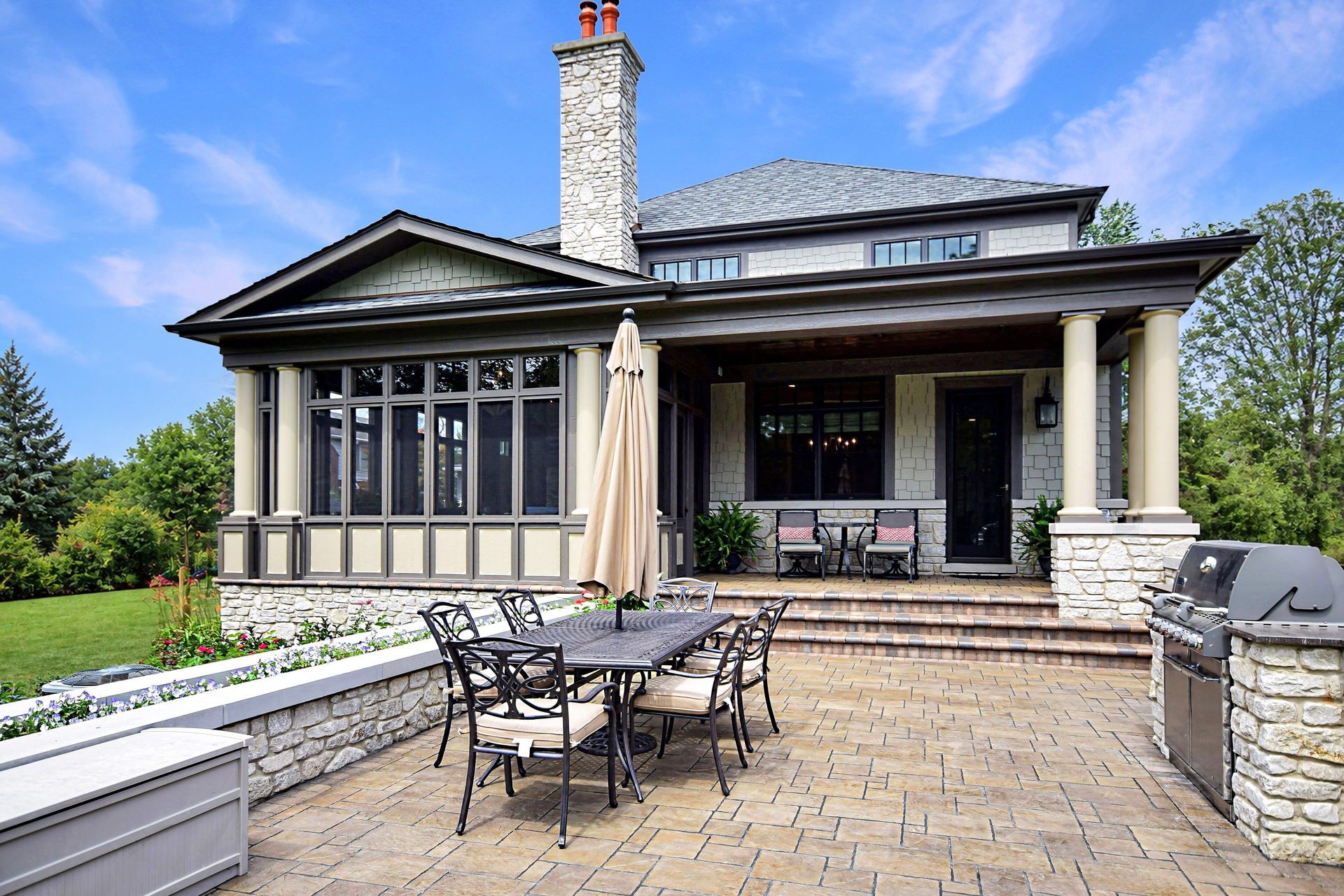 Patio dining area in front of a stone house with columns, patio furniture, and a closed umbrella.