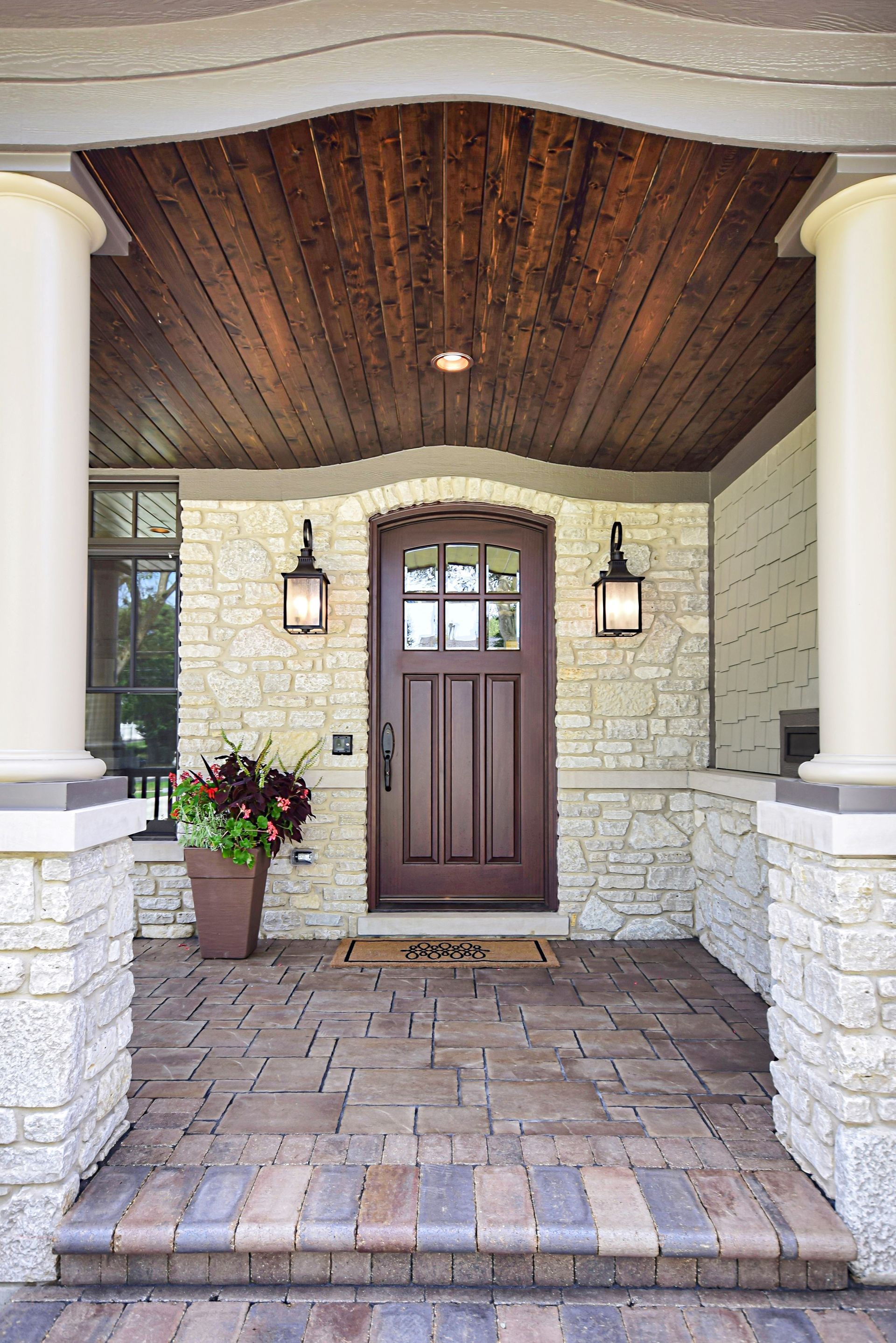 Front porch with a brown front door, stone walls, hanging lanterns, and potted flowers beneath a wood ceiling