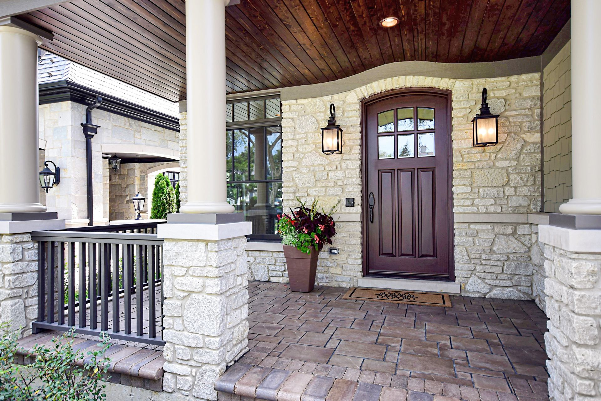 Front porch with stone columns, dark wood door, wall lanterns, and potted flowers under a wood ceiling
