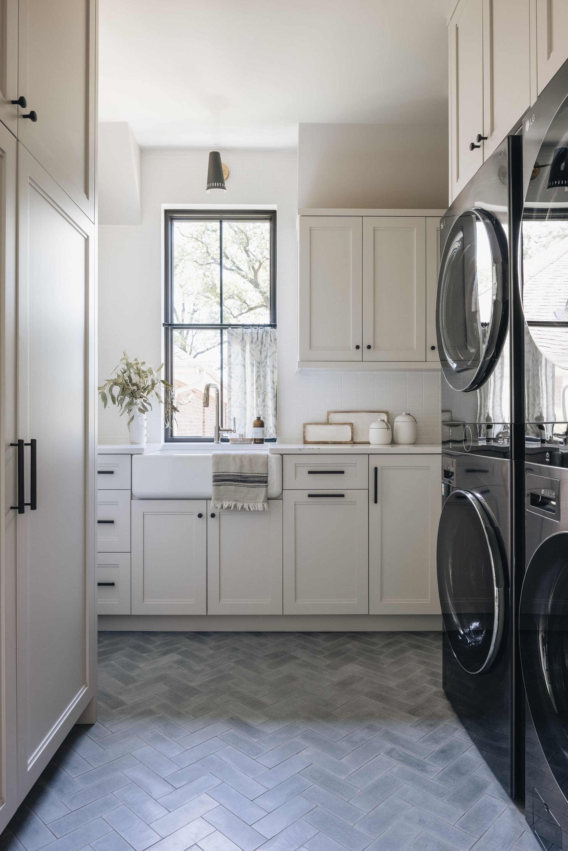 Bright laundry room with white cabinets, window, patterned floor, and stacked black washer-dryer.