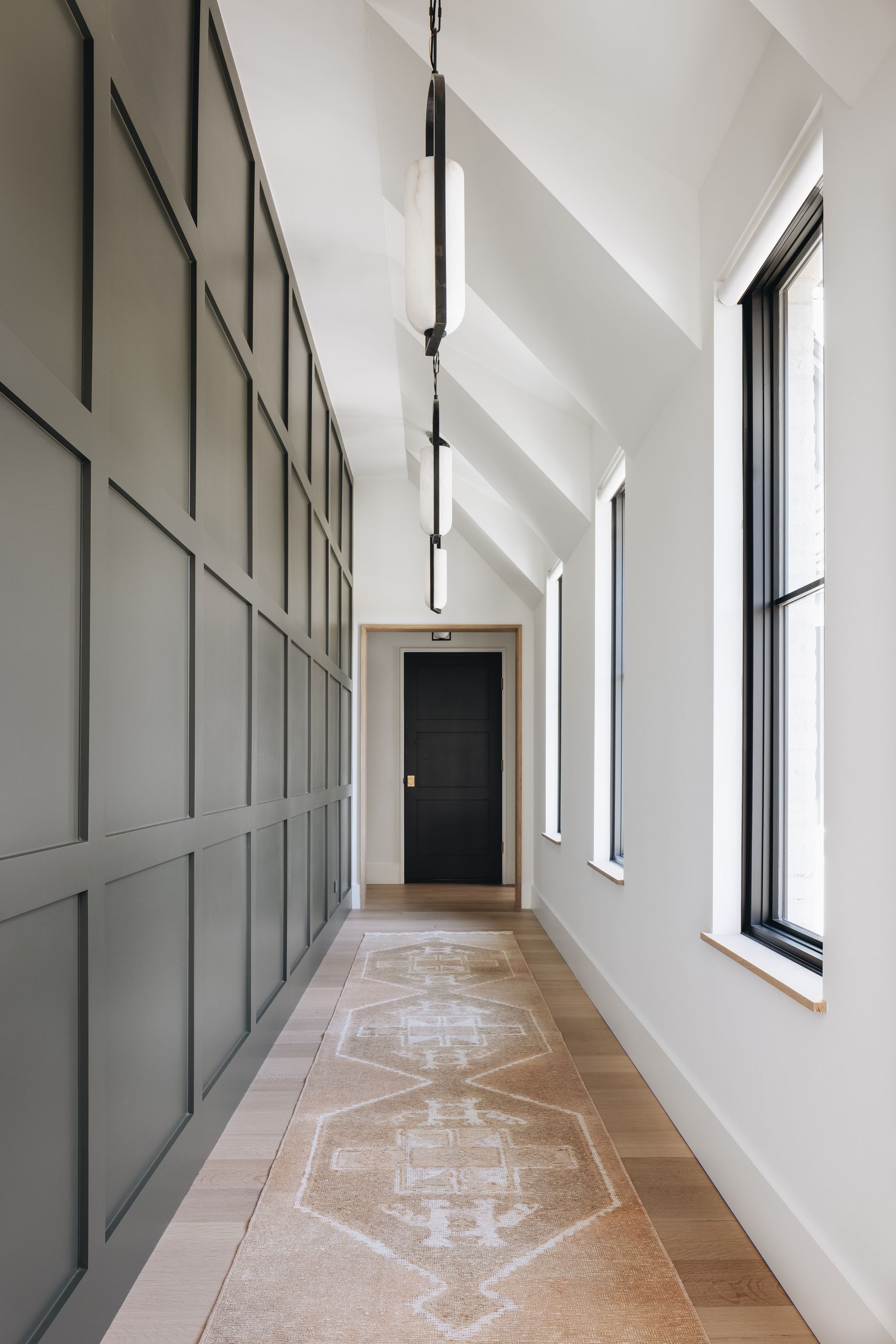 Modern hallway with tall windows, gray wall panels, white walls, and a dark door at the end.