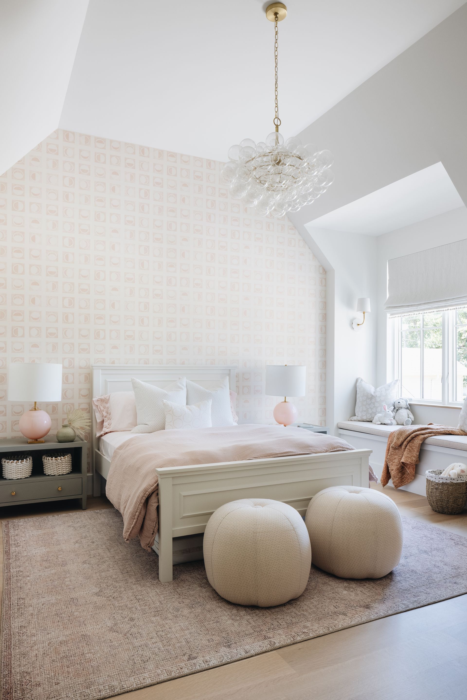 Bright bedroom with a pink bed, textured rug, two round poufs, and a crystal chandelier