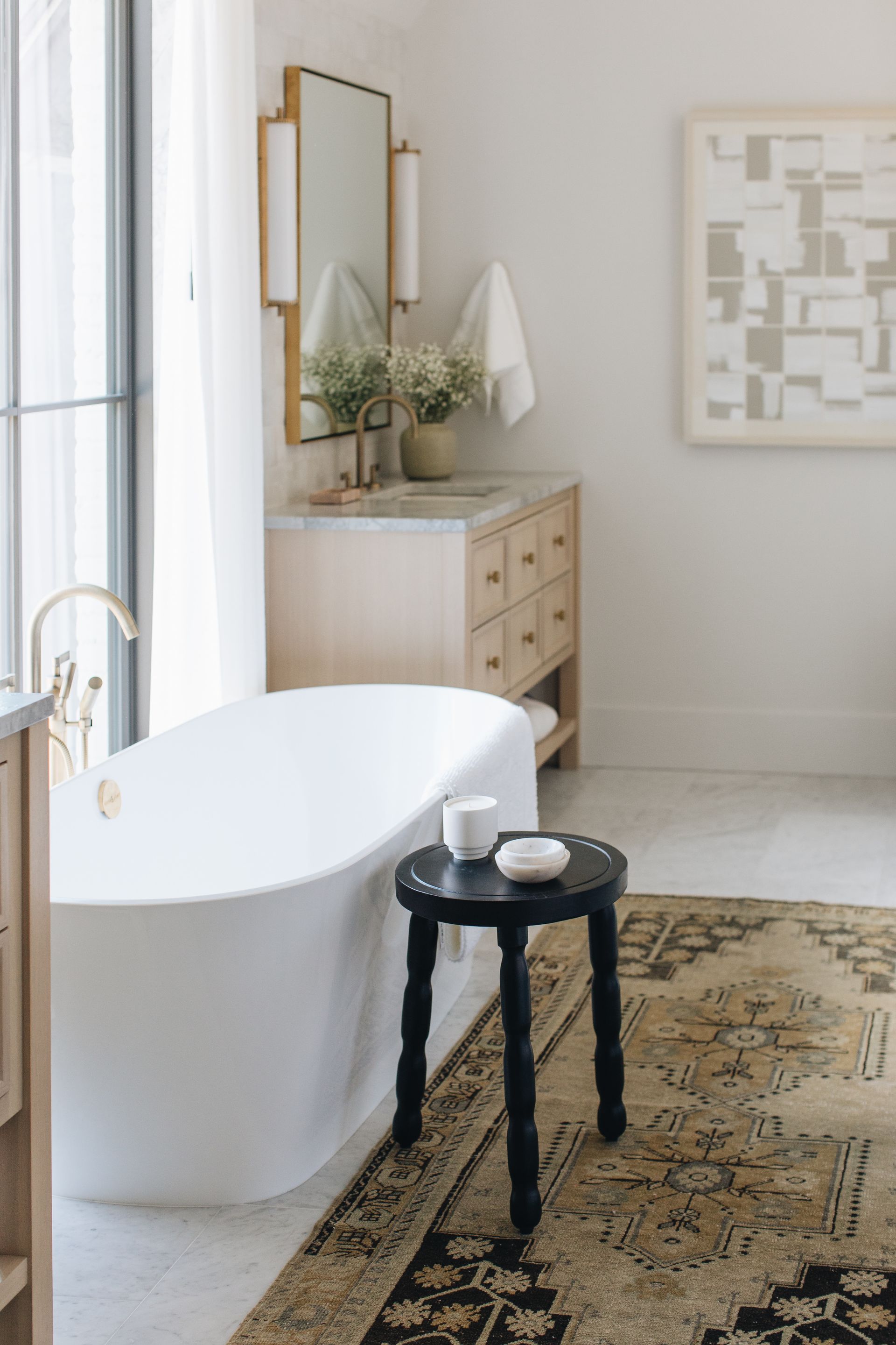 Bright bathroom with freestanding tub, black stool, patterned rug, and wooden vanity near a window.