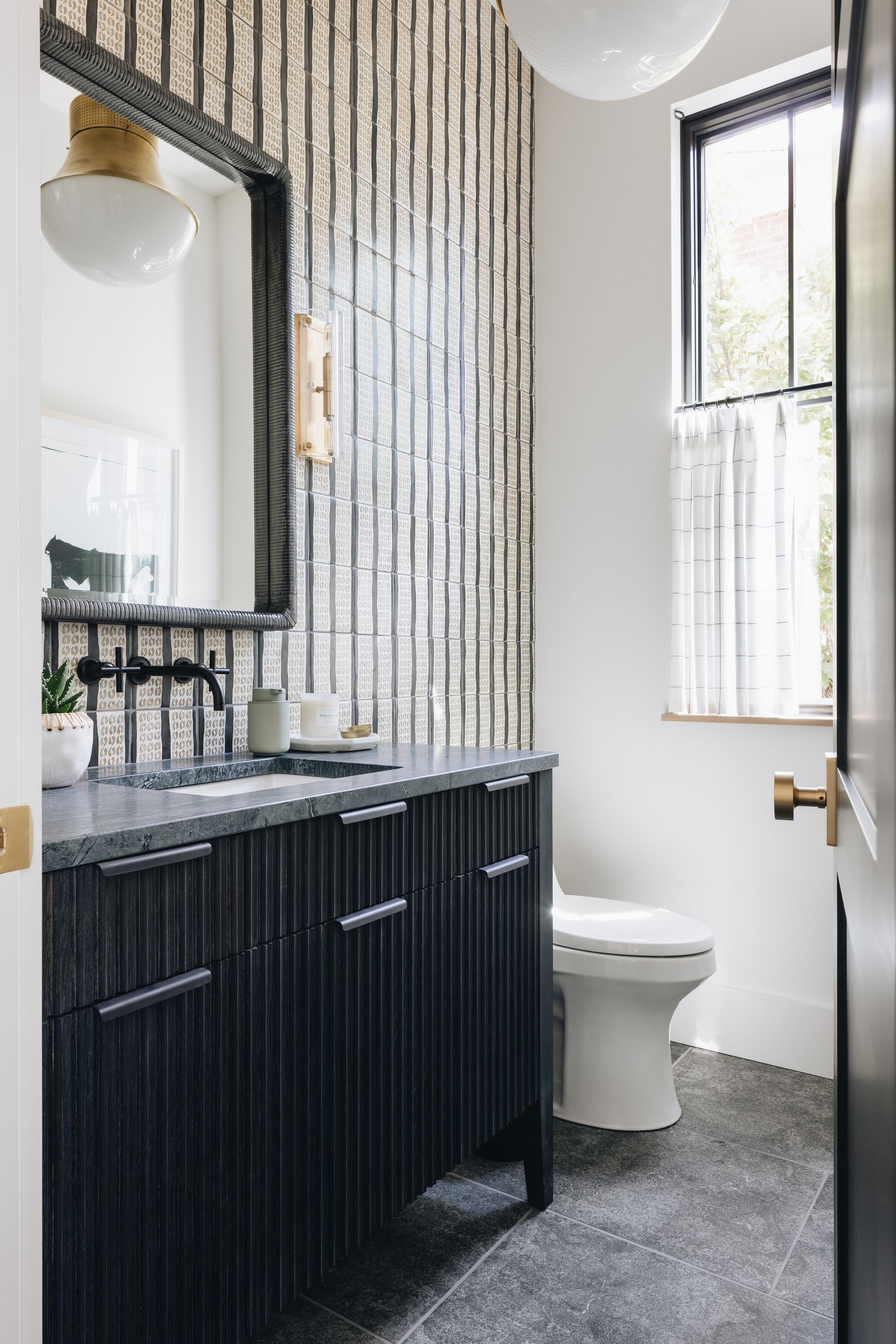 Modern bathroom with black vanity, round mirror, white toilet, and window with sheer curtain.