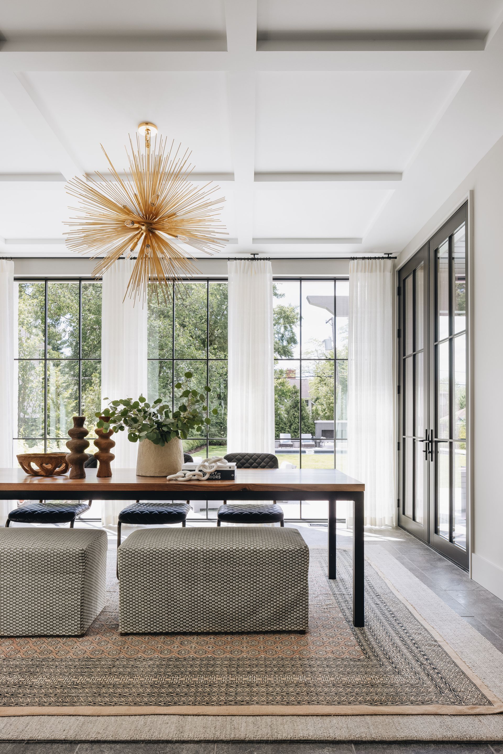 Bright modern dining room with a glass table, woven stools, potted plants, and a gold starburst chandelier.