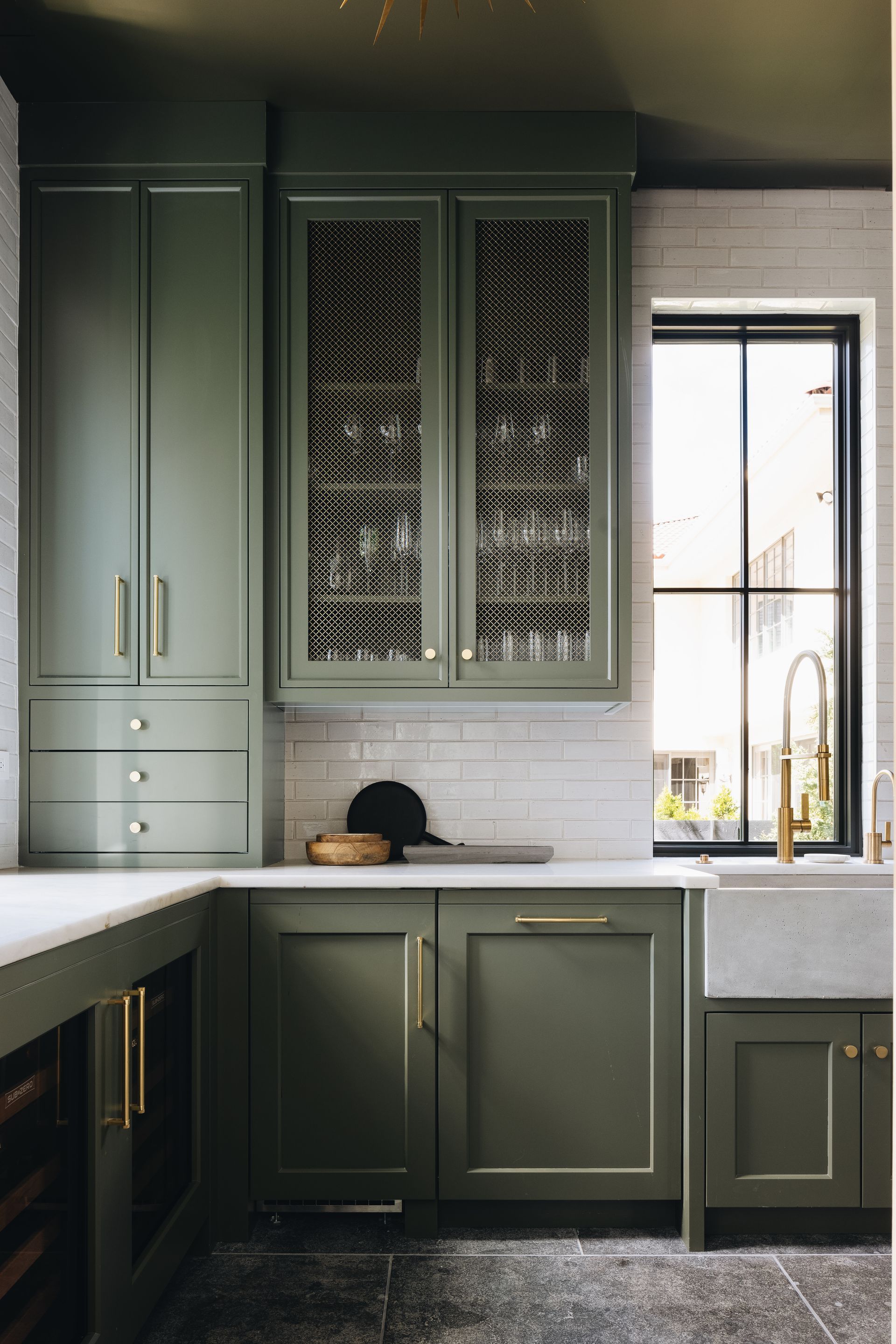 Green kitchen cabinets with brass pulls, gray countertop, and a bright window on the right