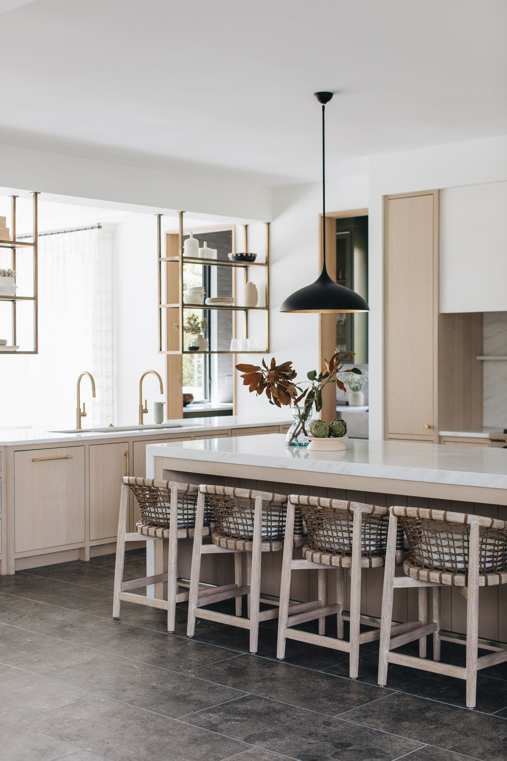 Bright kitchen with a large island, woven stools, black pendant light, and open shelving by a window.