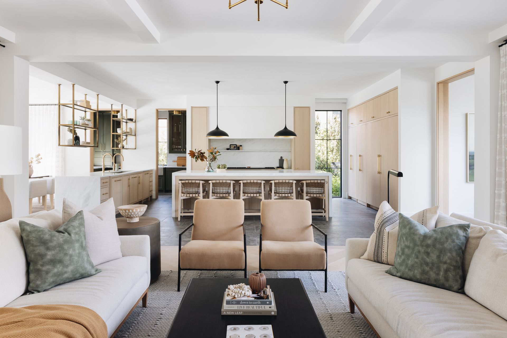 Bright open living room with beige sofas, two armchairs, black coffee table, and a kitchen in the background