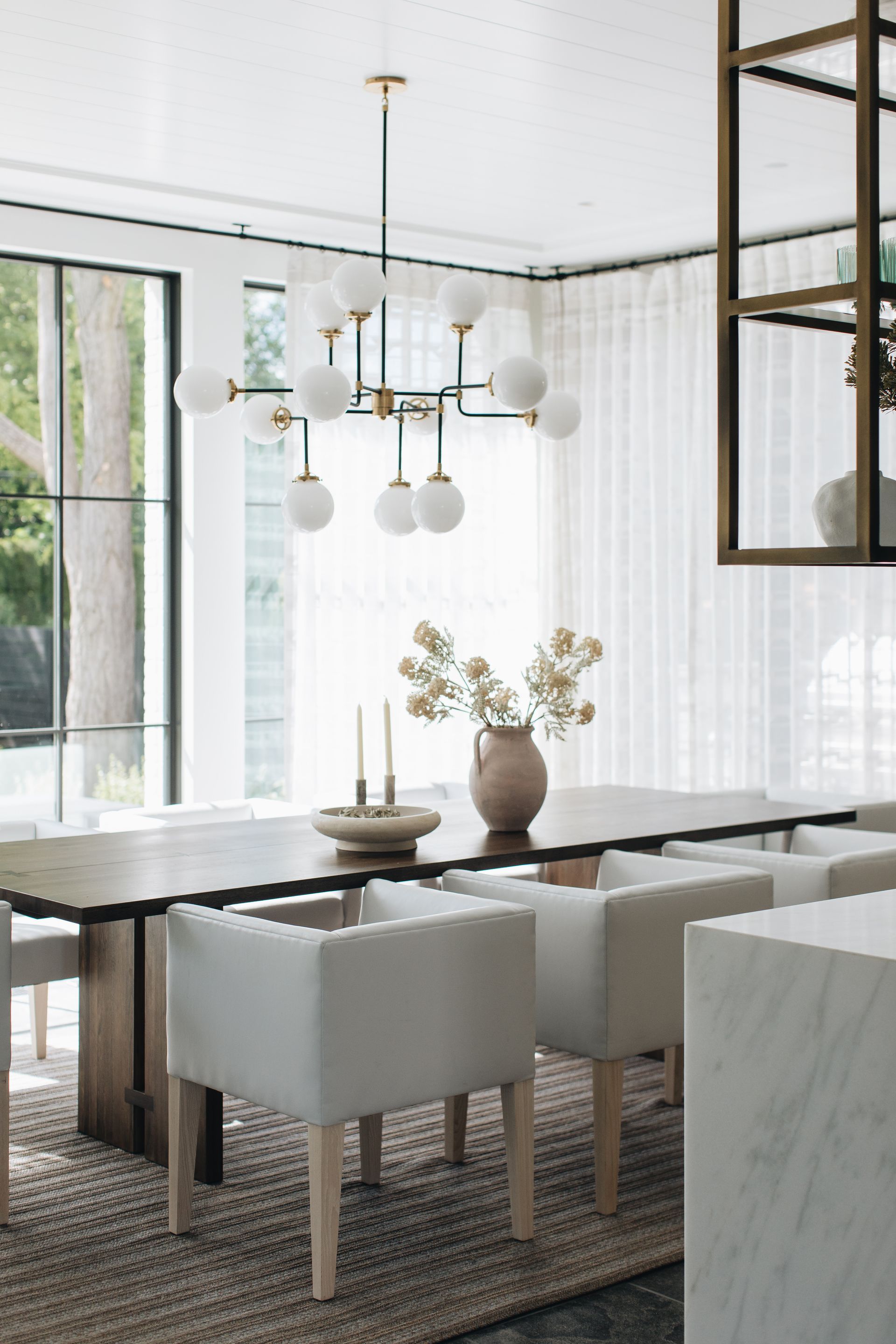 Bright dining room with a wooden table, white chairs, pendant lights, and large windows with sheer curtains