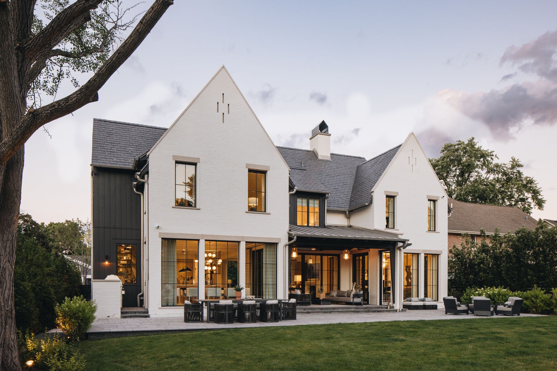 White two-story modern farmhouse with a dark roof, large windows, and a covered patio at dusk