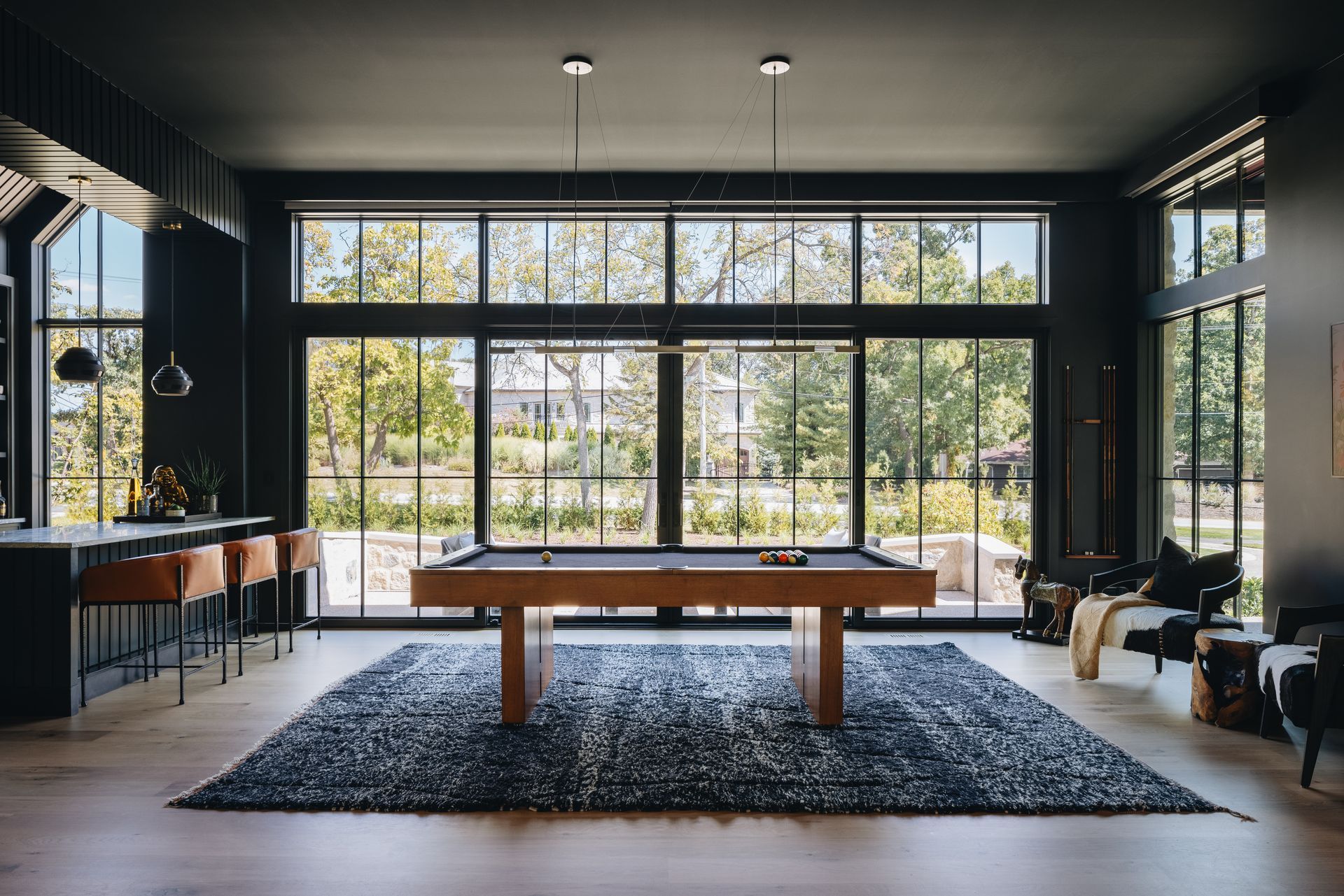 Modern living room with large windows, a pool table, dark walls, and sunlight streaming in
