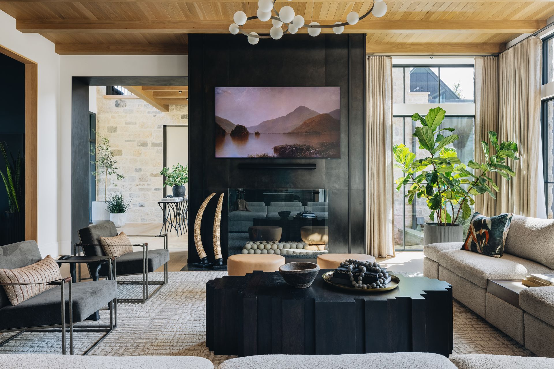 Modern living room with fireplace, wall TV, black coffee table, and large windows with plants