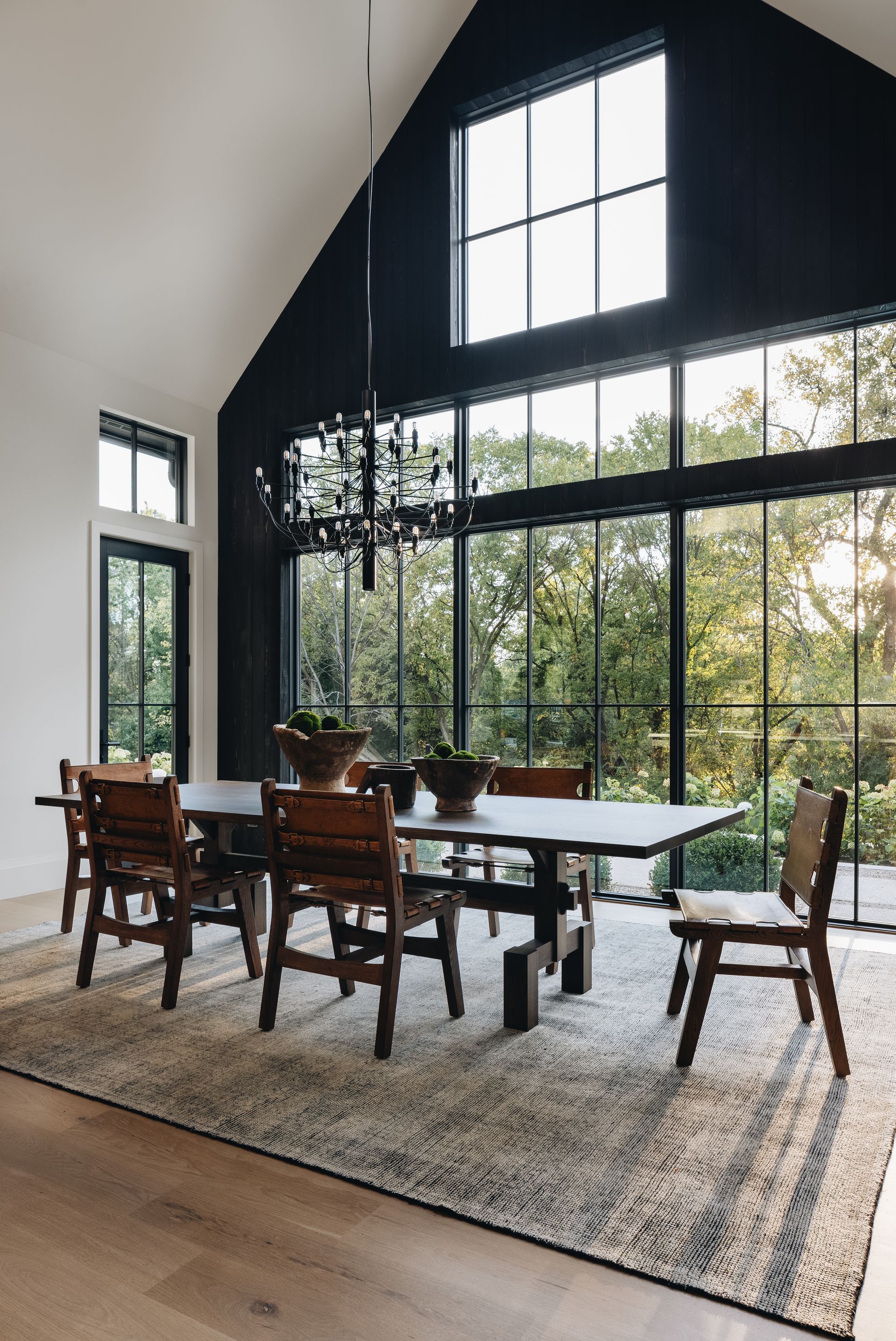 Modern dining room with tall black-framed windows, wooden table, chairs, and a large rug.