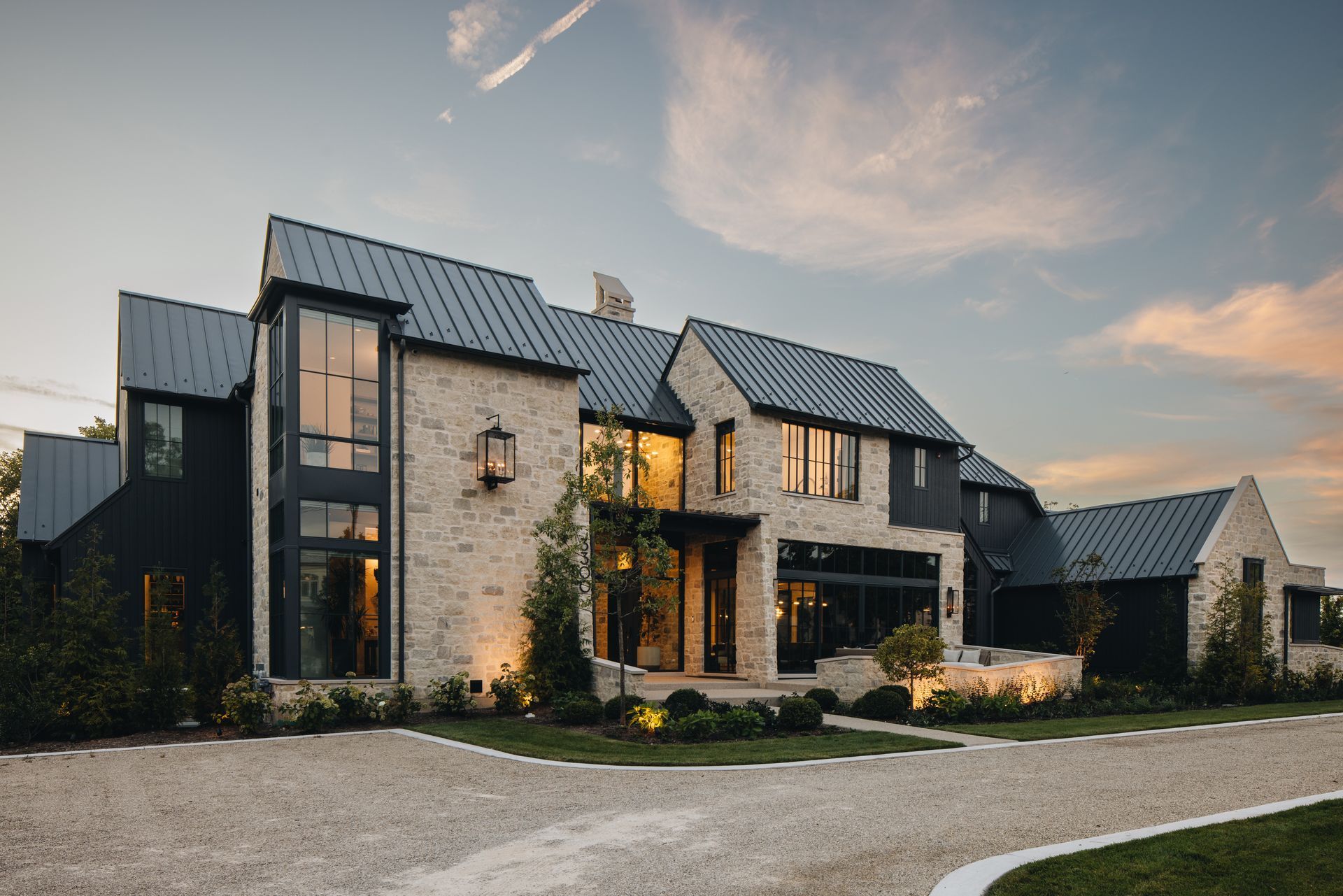 Modern stone house with dark metal roof, large windows, and a gravel driveway at sunset