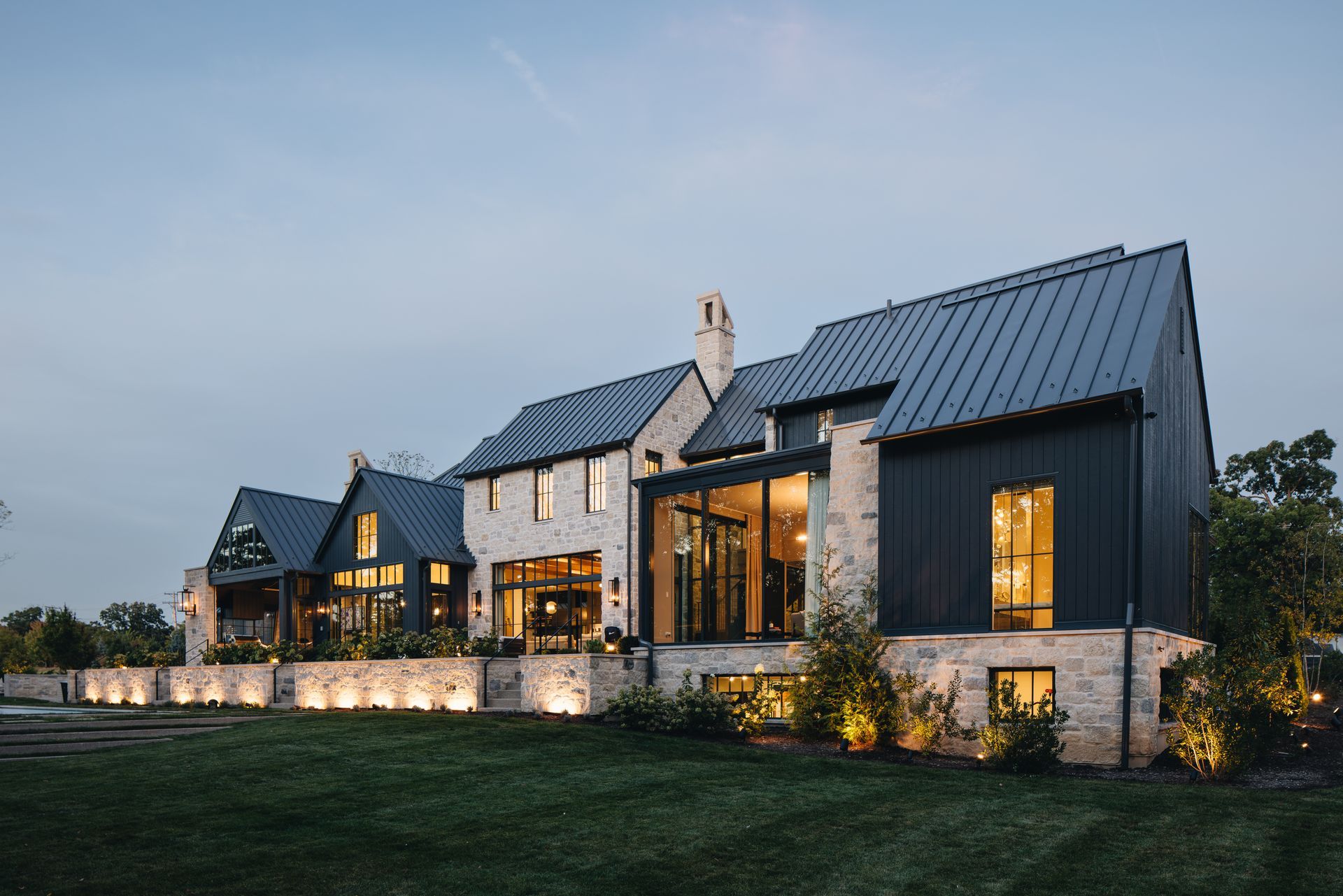 Modern stone house with dark metal roof and warm interior lights at dusk