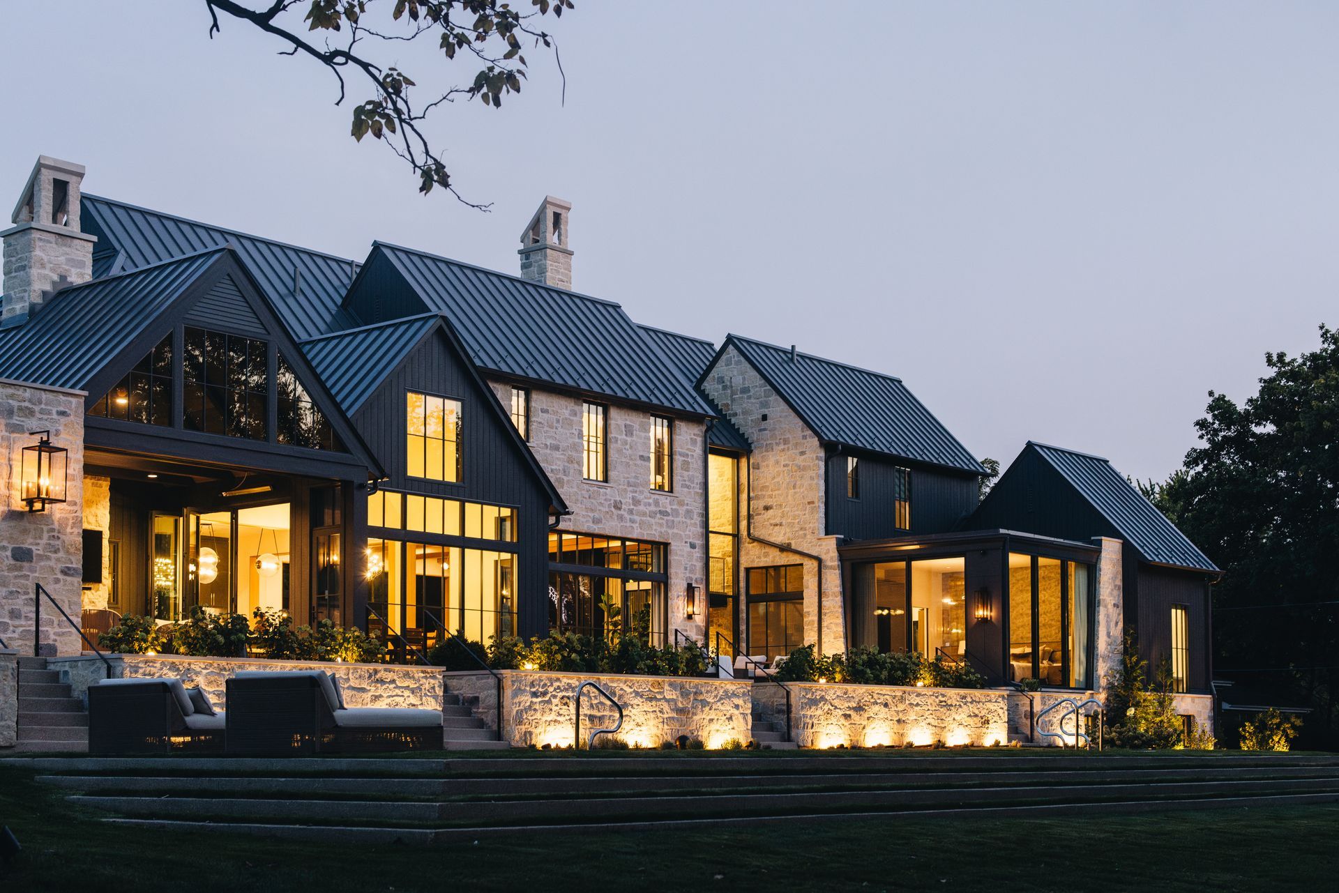 Large stone house with warm interior lights at dusk, featuring a dark roof, patio, and illuminated garden walls