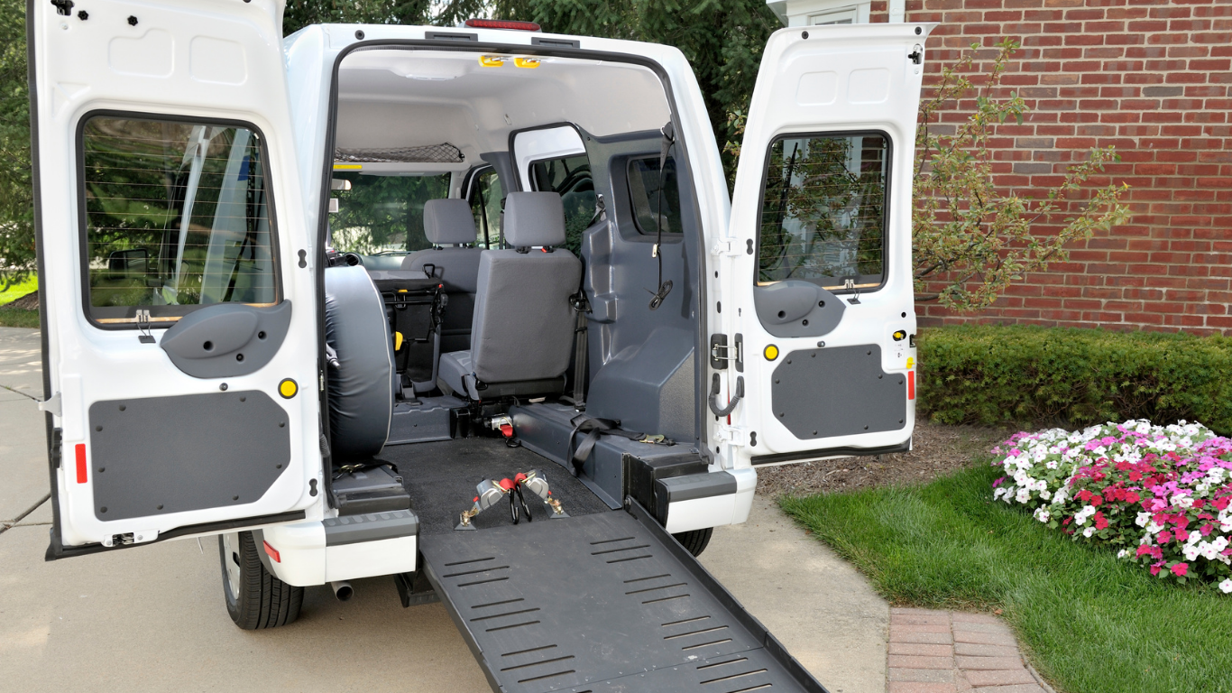 A white wheelchair-accessible van with rear doors open and a lowered access ramp extended onto a concrete driveway.